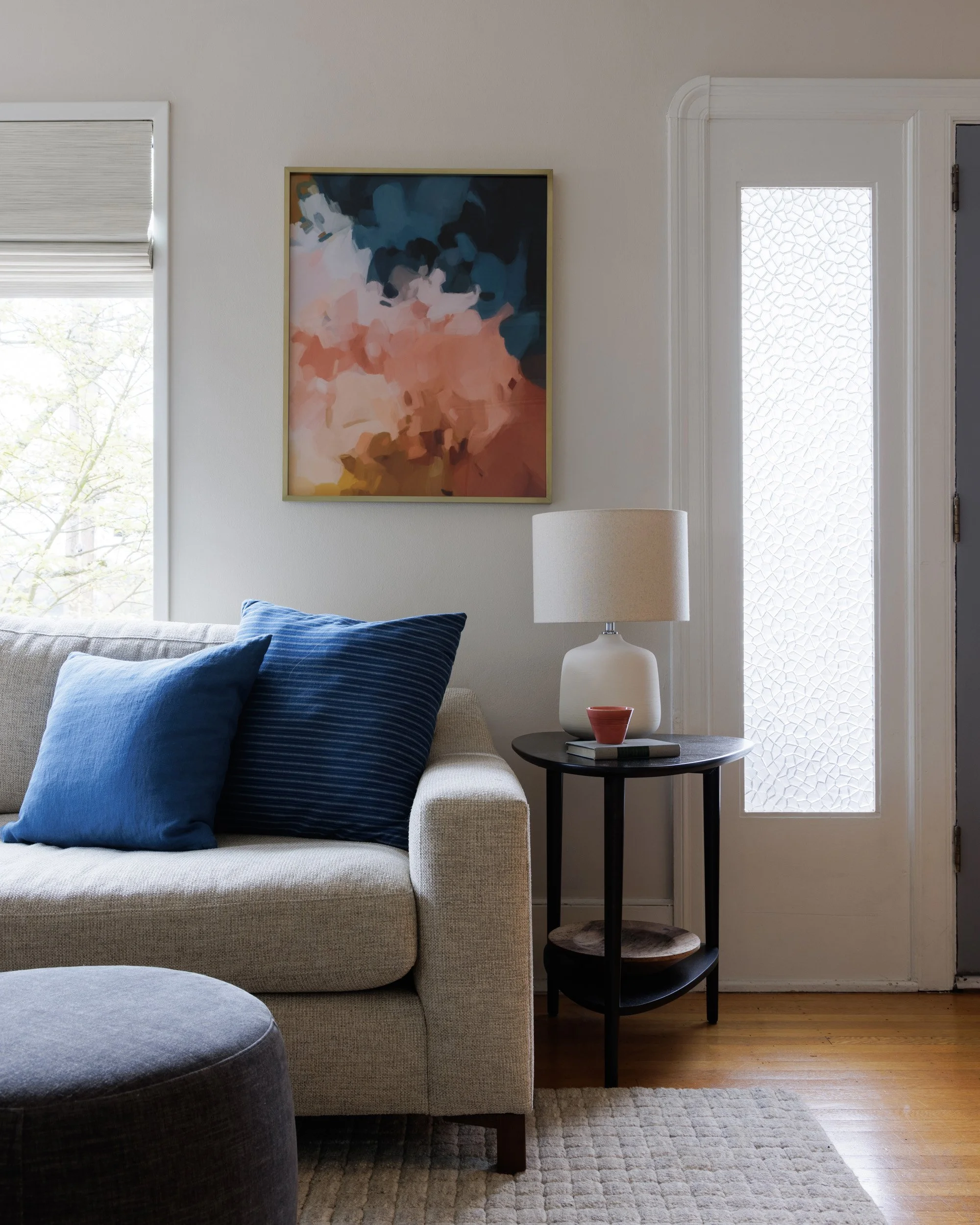 A transitional living room with a beige sofa and blue pillows, a side table with a white lamp, and a frosted glass window by Full Bloom Interior Design in Seattle.