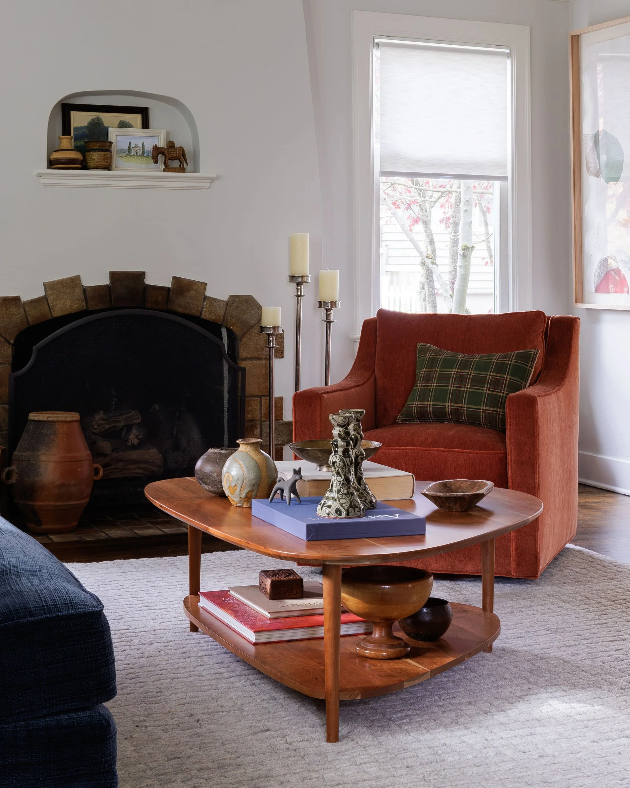 Tudor living room featuring a white stucco fireplace with a curated mantle of pottery and decor. A dark orange armchair with a plaid accent pillow sits behind a styled mid-century modern coffee table by Full Bloom Interior Design.