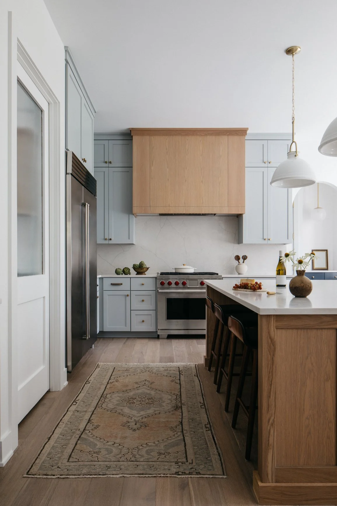 A transitional kitchen with light blue cabinets, a stainless steel refrigerator, a wooden range hood, stainless steel range, marble backsplash, and a white countertop island with bar stools, decorated with vases, a cheeseboard, flowers and wine.