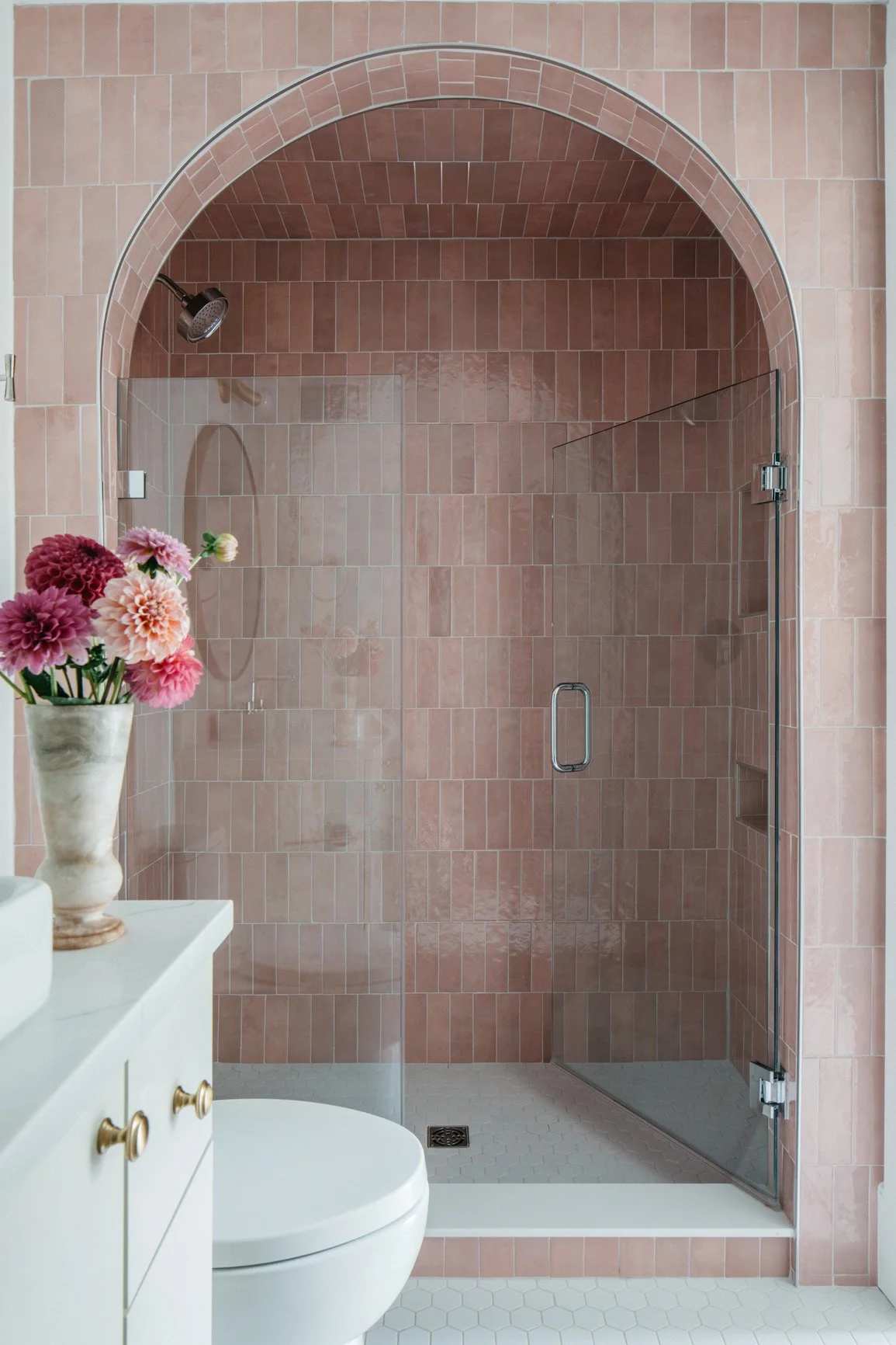 Bathroom with pink tiled shower area, glass door, white toilet, and a vase with pink flowers on a white vanity.