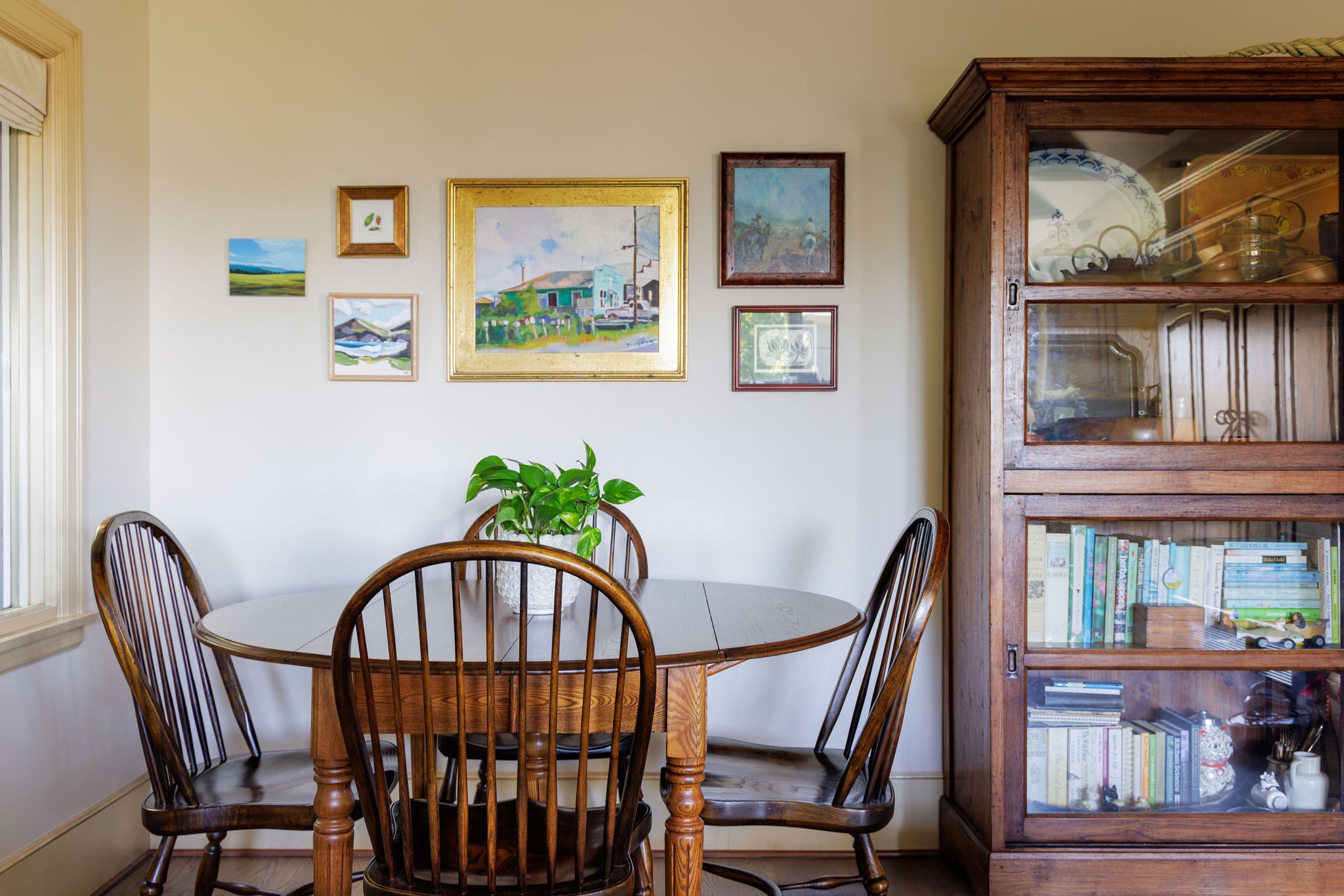 Breakfast nook featuring a large landscape gallery wall in gold frames, a round oak table, and vintage spindle chairs by Full Bloom Interior Design in Seattle.