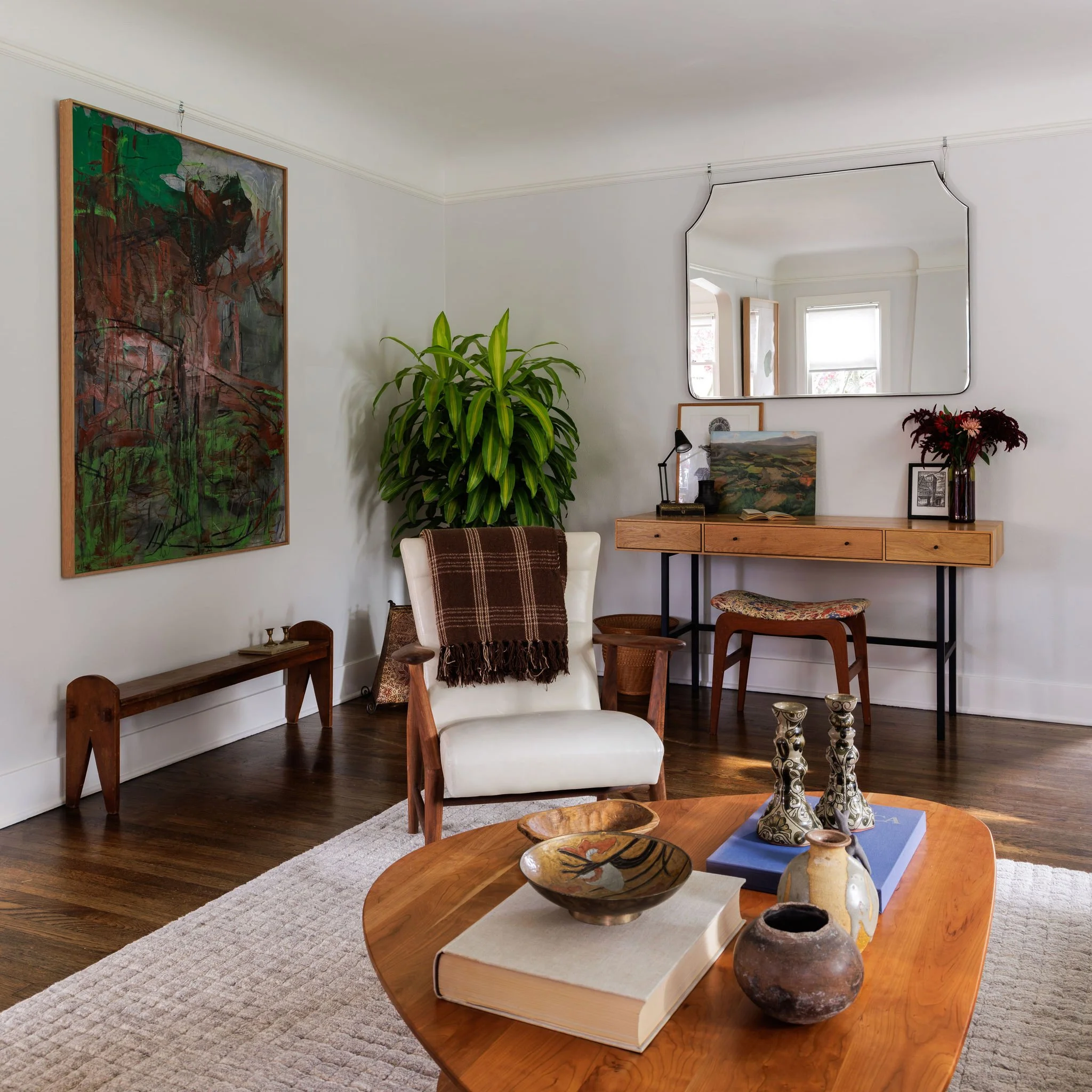 Mid-century modern living room corner with a white armchair, wooden desk with mirror hanging above, wall art and large indoor potted plant by Full Bloom Interior Design in Seattle.