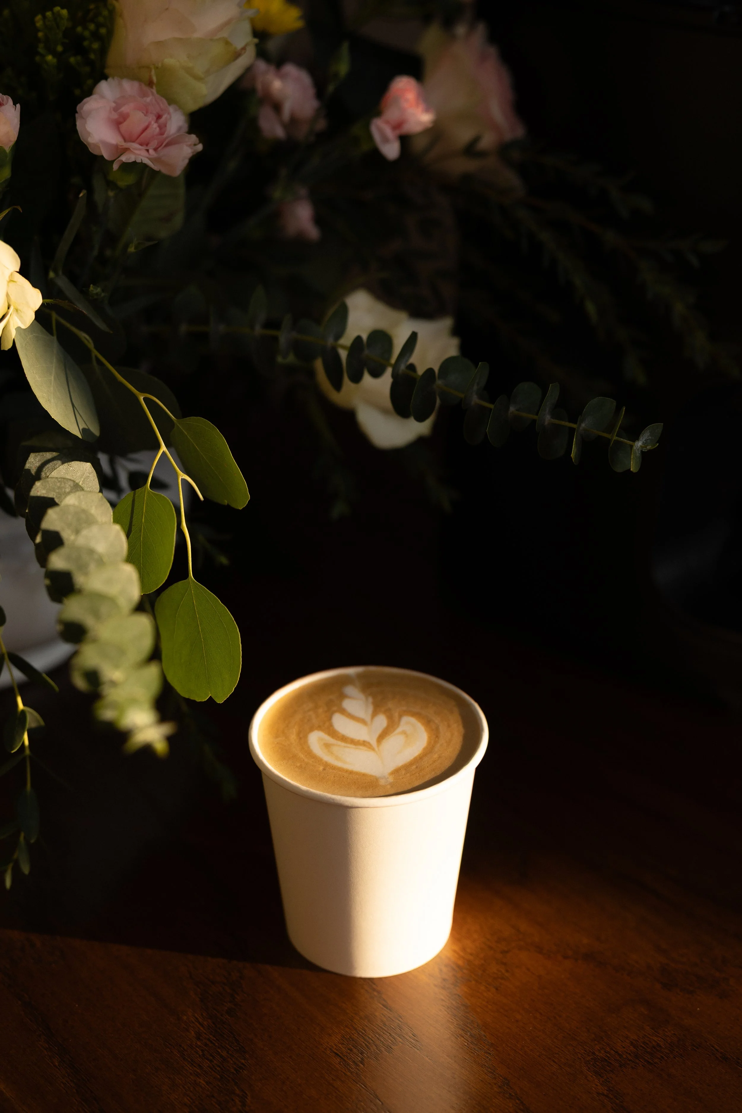 A cup of coffee with latte art sits on a wooden surface, illuminated by warm light, with green leaves and pink and white flowers in the background.