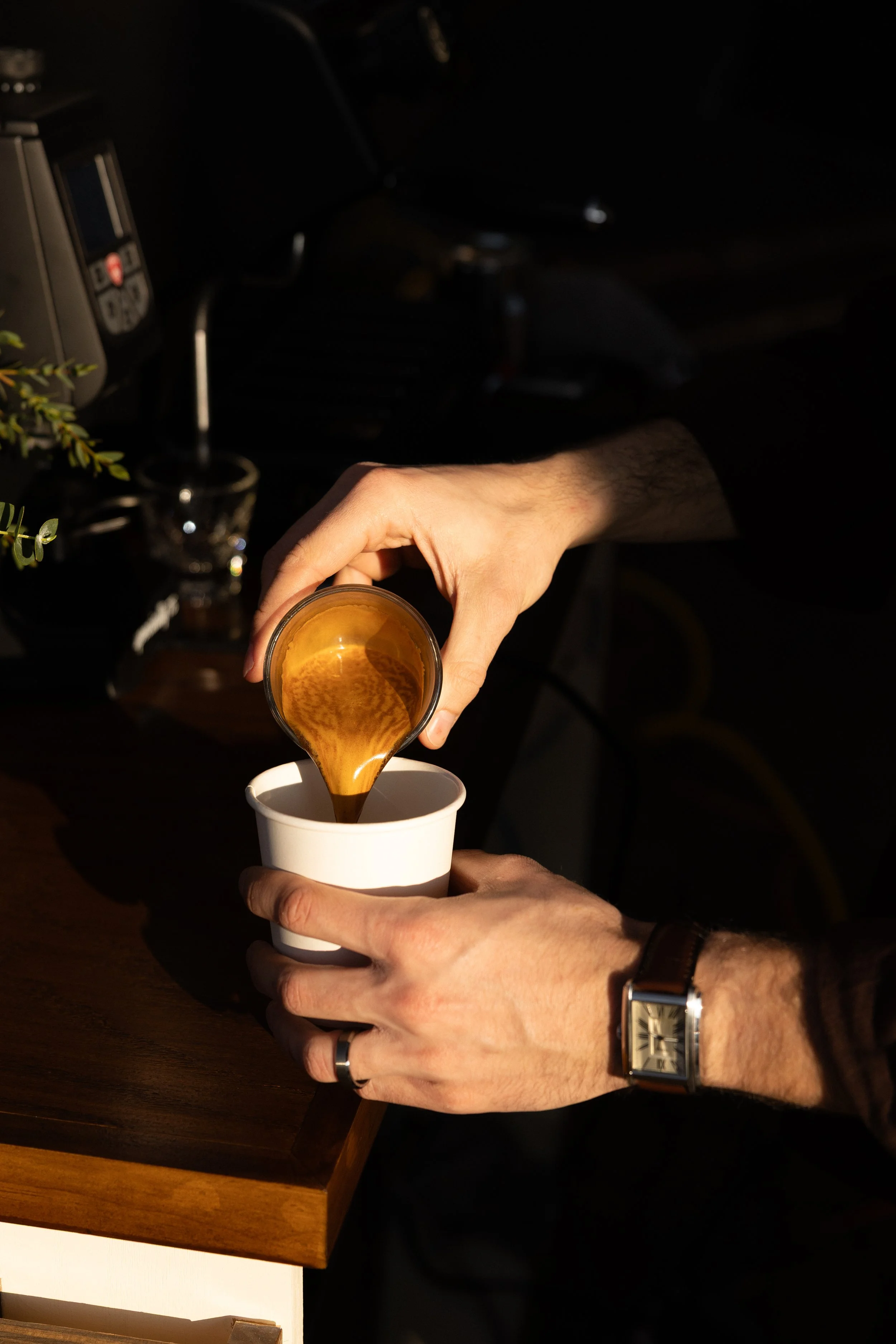 A person pours coffee from a small glass container into a white paper cup, with a watch on their wrist and a ring on their finger, on a wooden surface with a dark background.