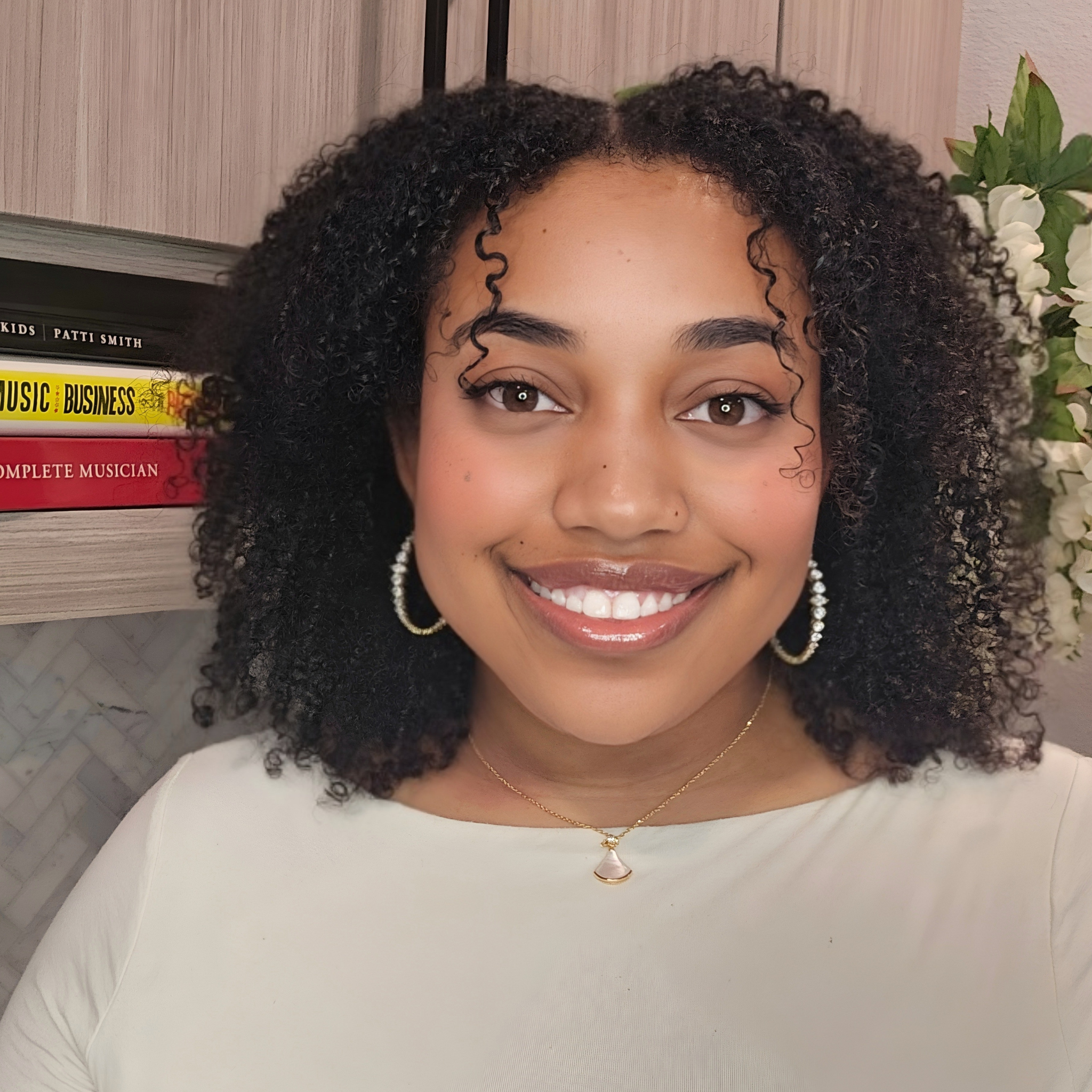 Smiling woman with curly black hair, wearing hoop earrings, a necklace, and a white top, standing in front of a bookshelf and a plant with white flowers.