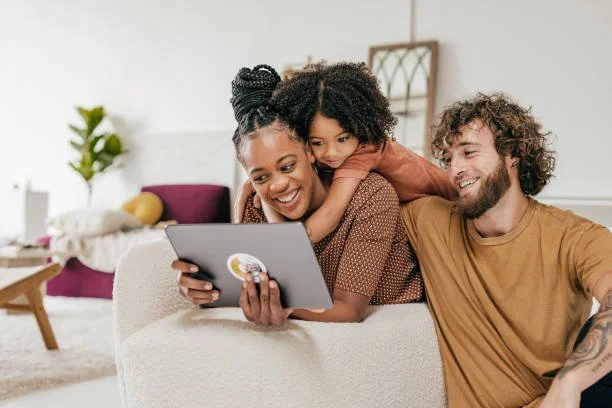 A happy family of three, an African American woman, a girl, and a Caucasian man, sitting on a white couch and looking at a tablet together in a bright living room.