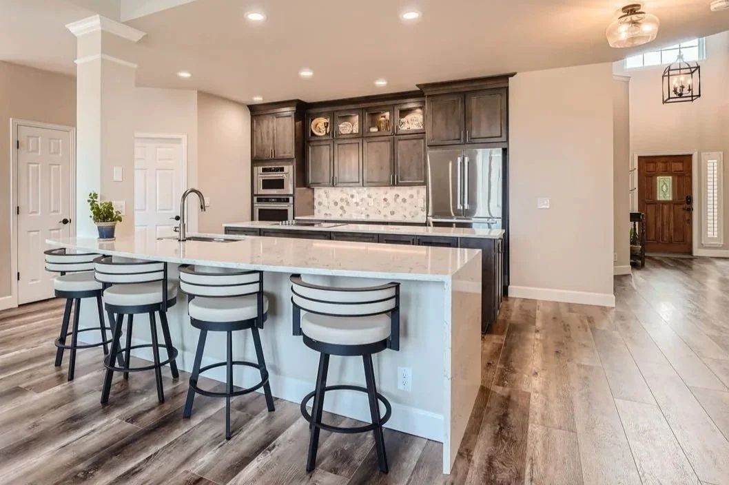 Modern kitchen with a large island, white countertops, dark wood cabinets, stainless steel appliances, and six white and black barstools.