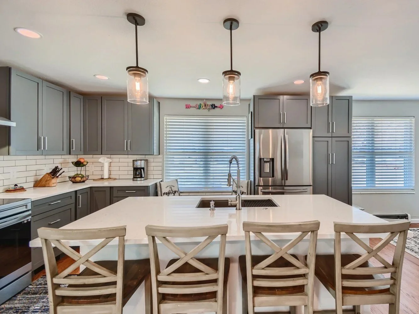 Modern kitchen with gray cabinets, white countertops, stainless steel refrigerator, island with a sink and four wooden chairs, pendant lights, and window blinds.