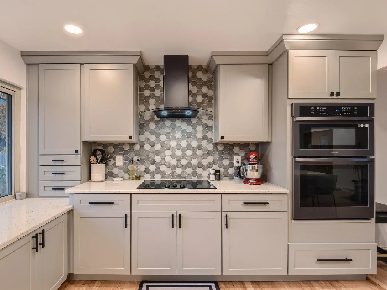 Modern kitchen with light gray cabinets, a hexagonal tile backsplash, built-in microwave and oven, a stovetop, a black range hood, and small kitchen appliances on the countertop.
