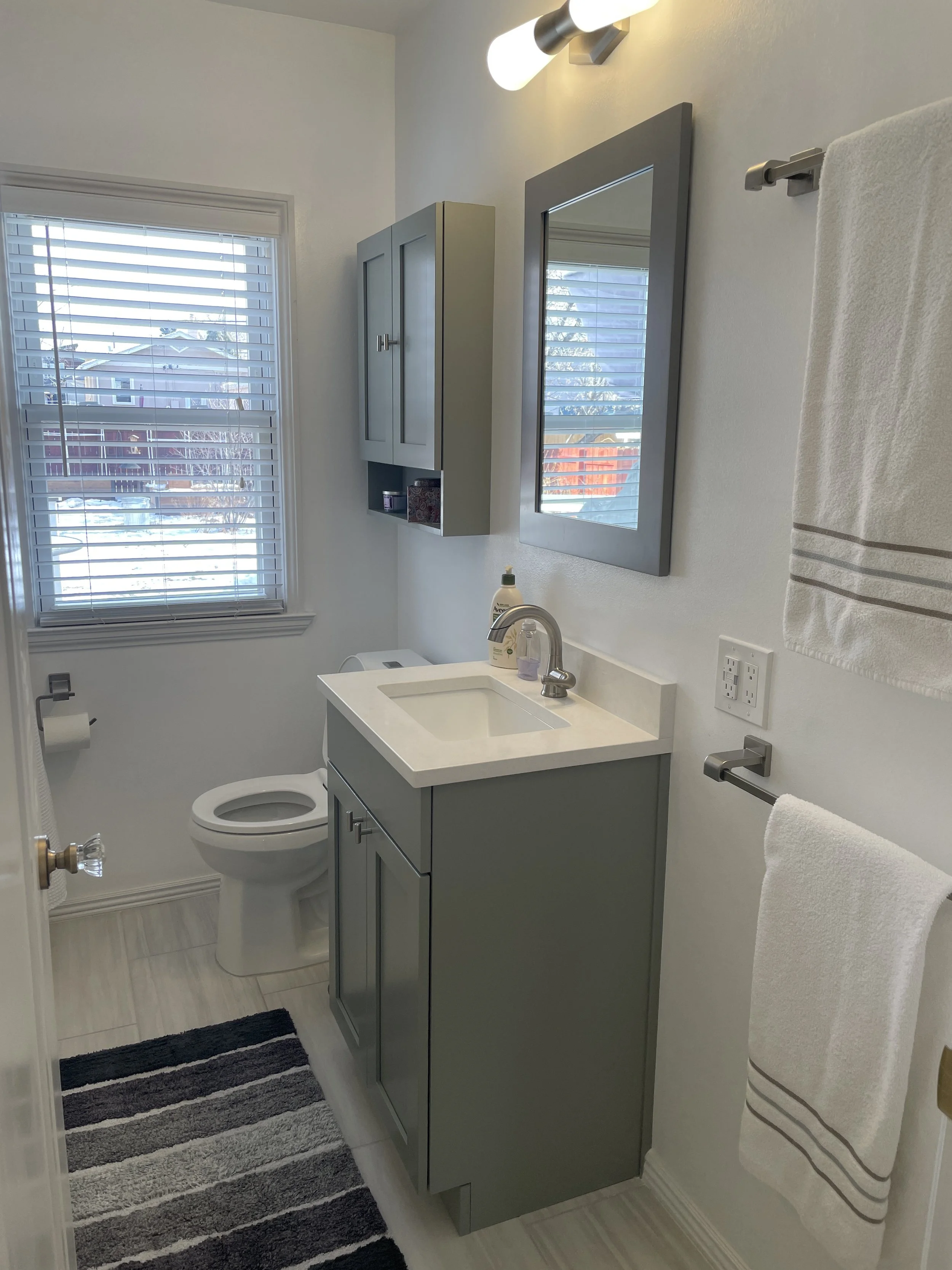 A small bathroom with a window, a toilet, a green vanity with a sink, a mirror, a towel rack with a white towel, and a cabinet above the toilet.