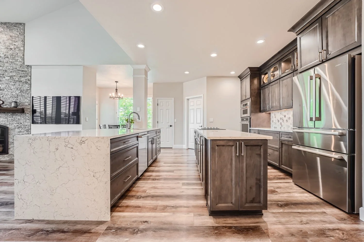 Modern kitchen with dark wood cabinets, stainless steel appliances, white countertops, wood flooring, and an open layout leading to a dining area with a chandelier.
