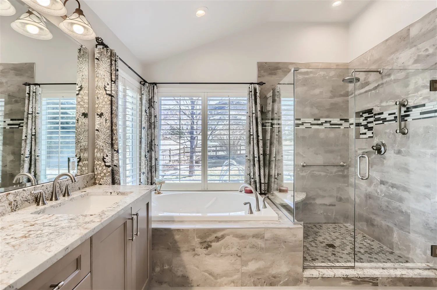 Modern bathroom with a large window, a bathtub, and a shower area with glass doors, gray tile walls, a granite countertop, and floral curtains.