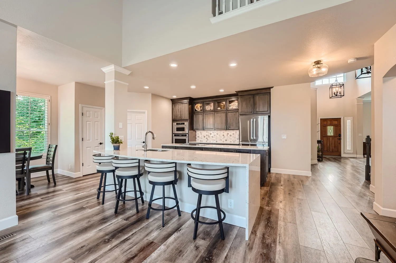 Open kitchen with white island, dark wooden cabinets, stainless steel appliances, and wood flooring, with natural light from windows and ceiling lights.