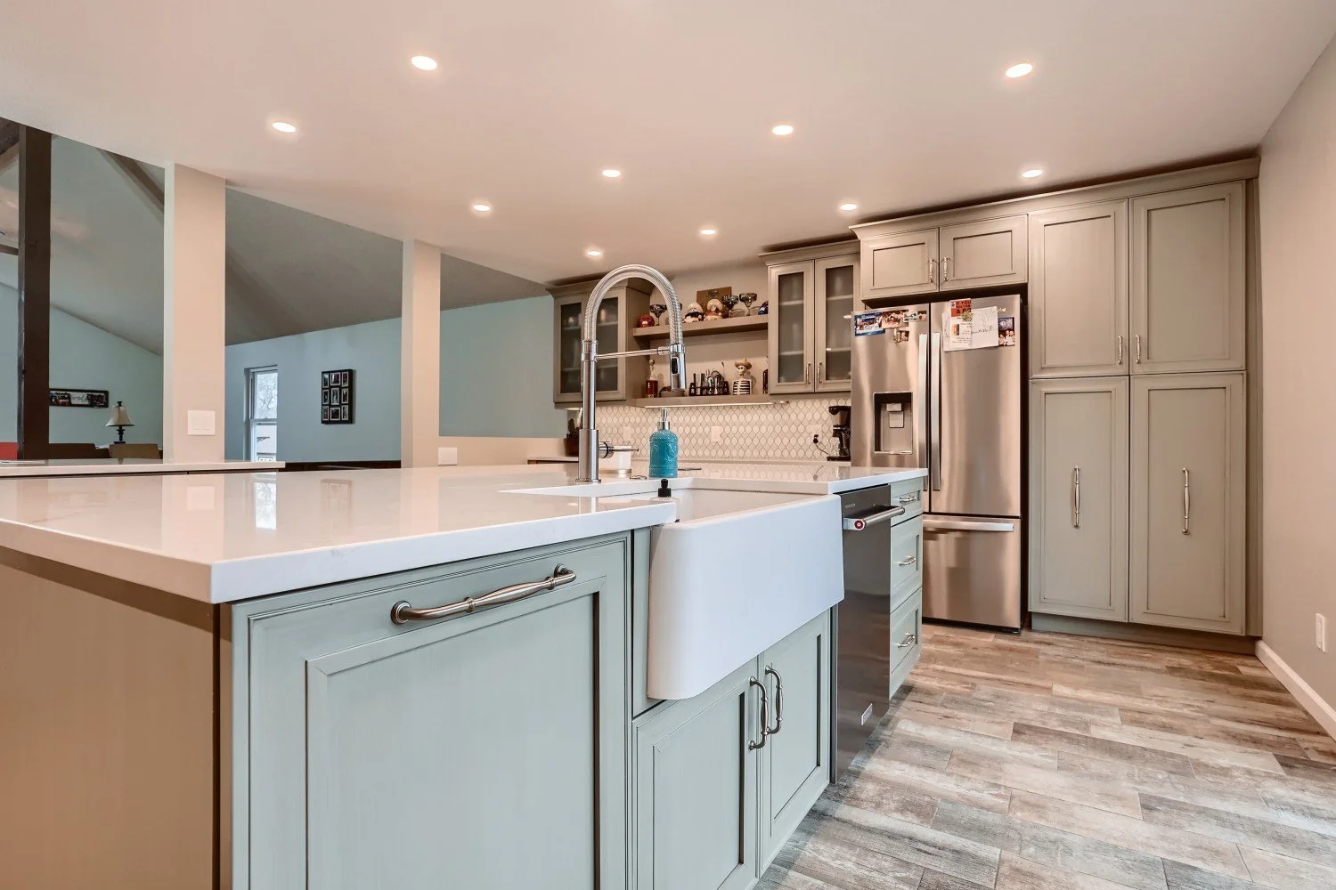 Modern kitchen with light green cabinets, white countertop island, stainless steel refrigerator, farmhouse sink, and wooden flooring.