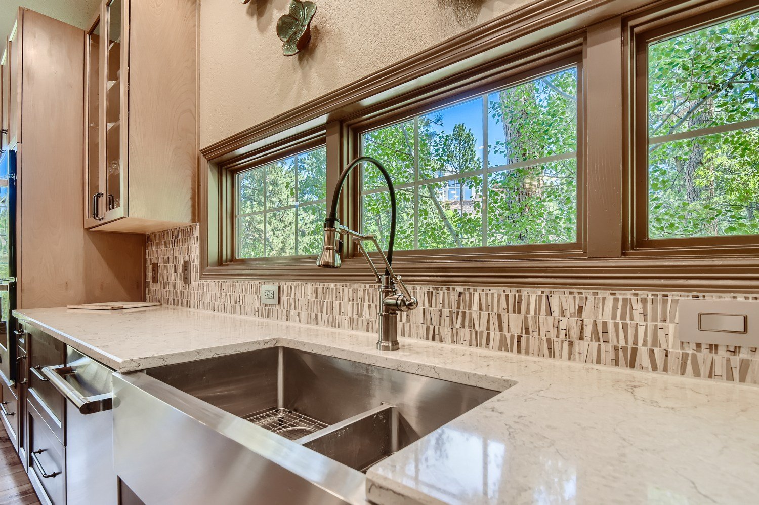 Kitchen sink with a modern faucet, cream-colored countertop, mosaic tile backsplash, wooden cabinets, and large windows with trees outside