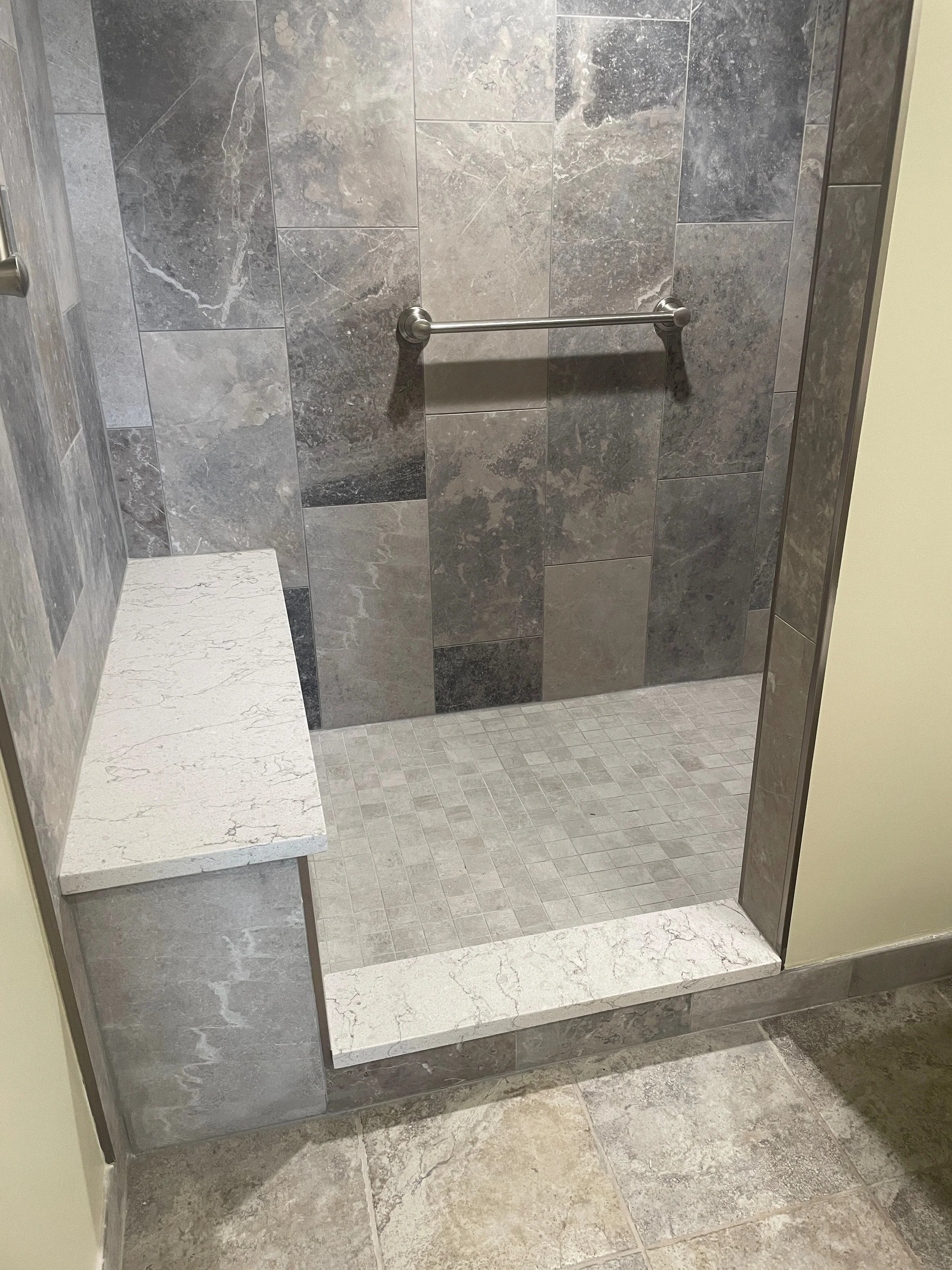 A bathroom with a walk-in shower featuring gray tiles and a bench, a white toilet with the seat covered in plastic, beige walls, and tiled flooring.