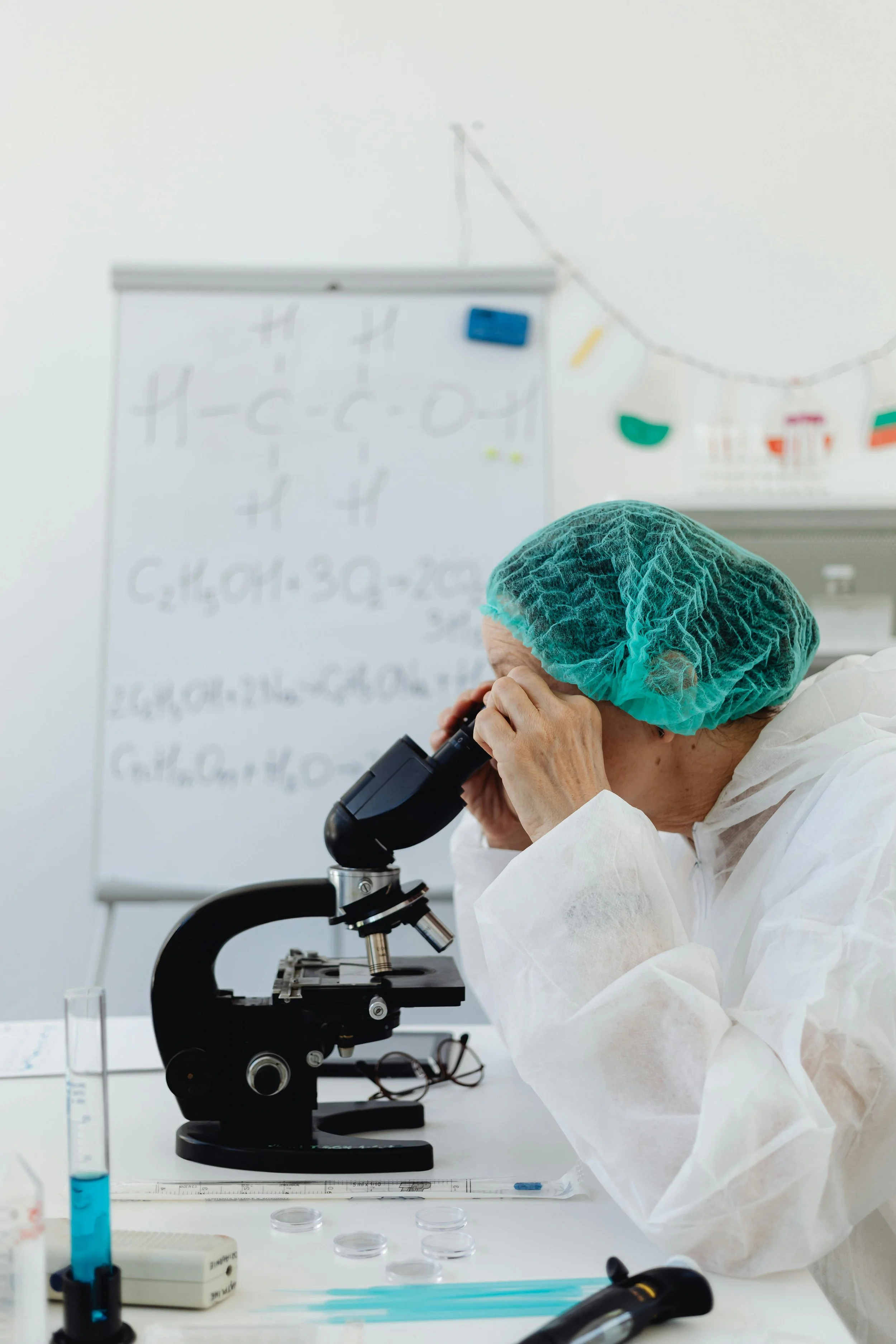 A scientist in a white lab coat and blue hair cover, using a microscope in a laboratory with scientific equipment and a whiteboard with chemical formulas in the background.