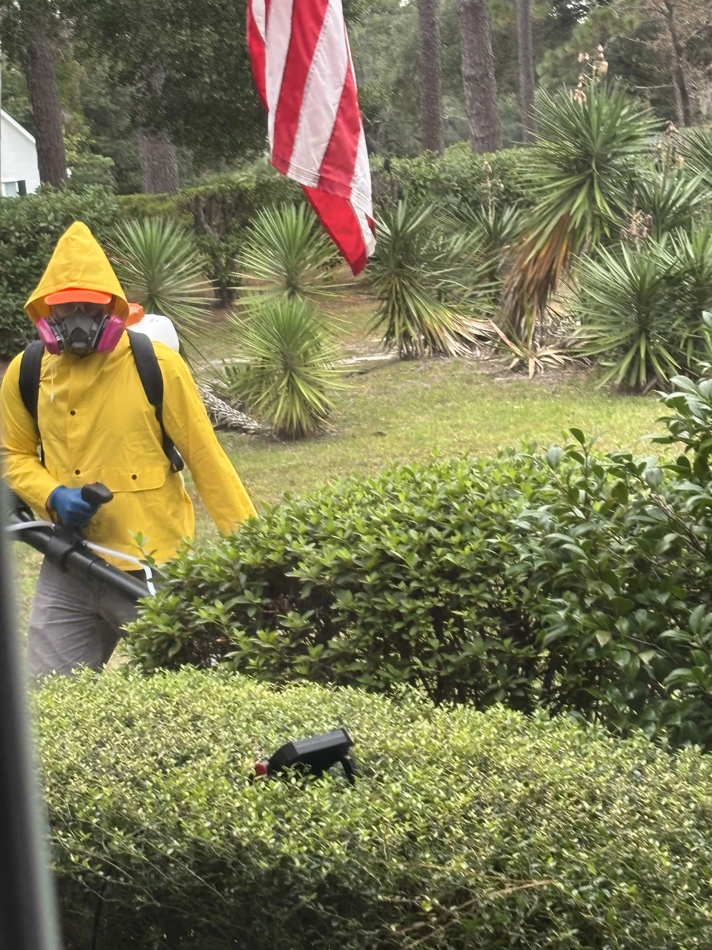 Person in a yellow raincoat and protective gear operating yard equipment outdoors with bushes and trees.