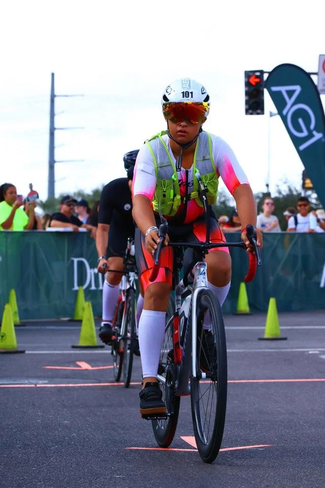 A young cyclist wearing a helmet, sunglasses, colorful cycling jersey, and a reflective safety vest crossing a line on a race track with spectators in the background.