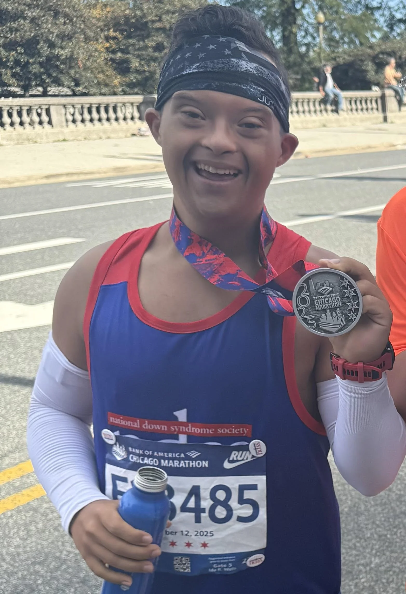 A smiling marathon runner holding a finisher medal at the Chicago Marathon, wearing a blue and red race shirt, a black star-patterned headband, and a red watch, with a race bib number 5485.