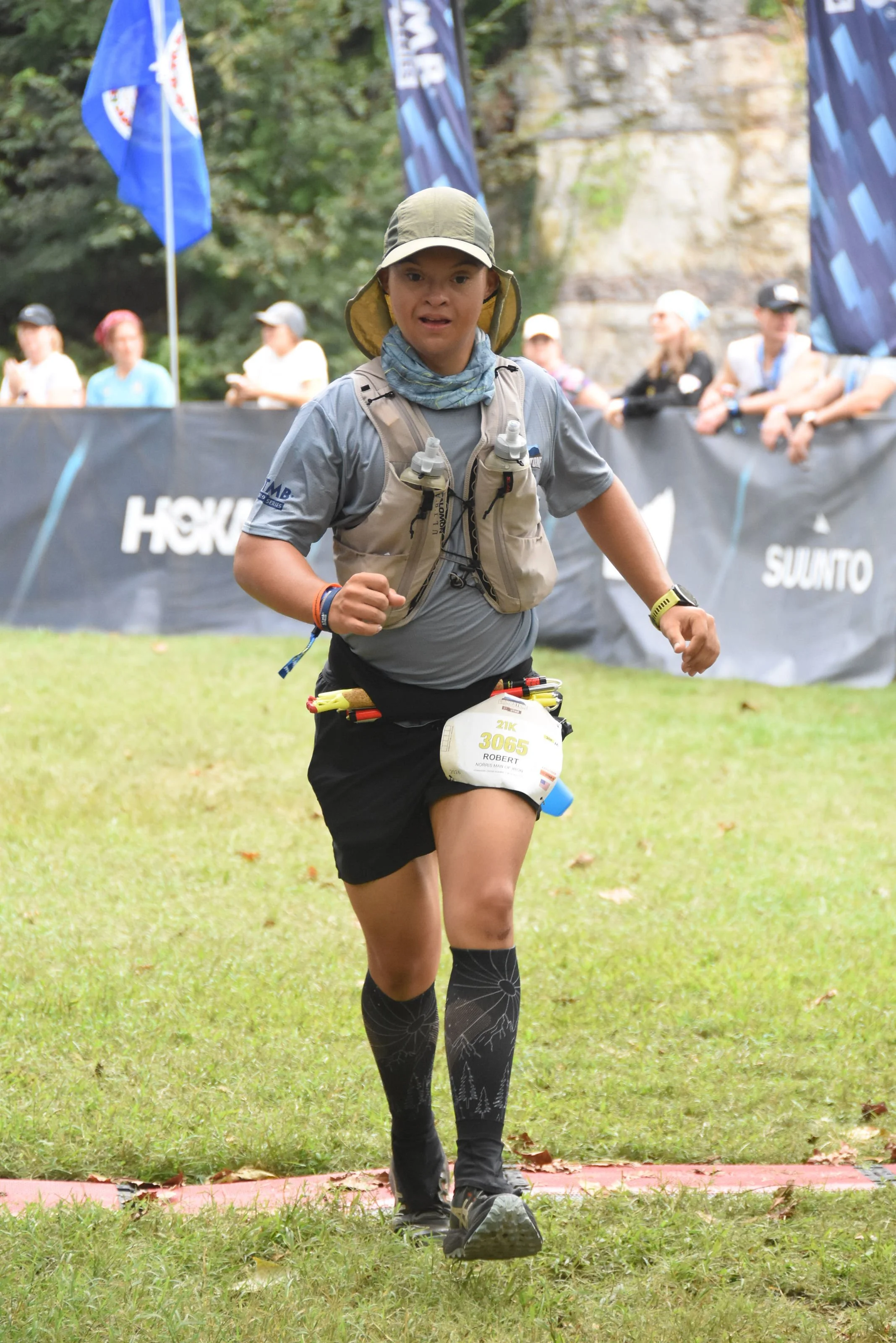 A young male athlete running in a race, wearing a light gray shirt, black shorts, patterned black knee-high compression socks, a beige hydration vest, a beige cap, and a race bib with the number 3065 and the name Robert. He is outdoors on grass, with a black barrier and spectators in the background.