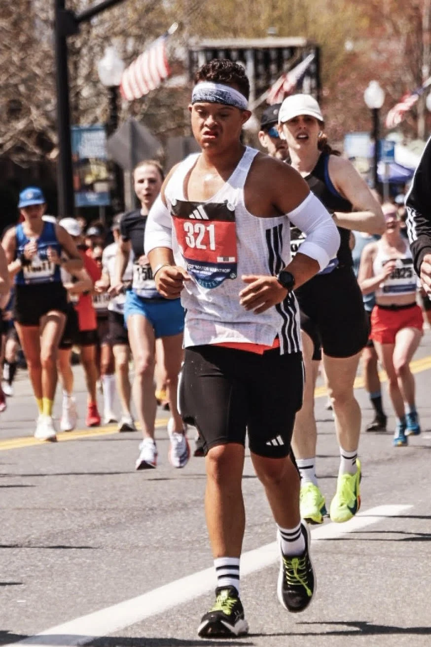 A group of runners participating in a road race on a sunny day, with flags and spectators in the background.