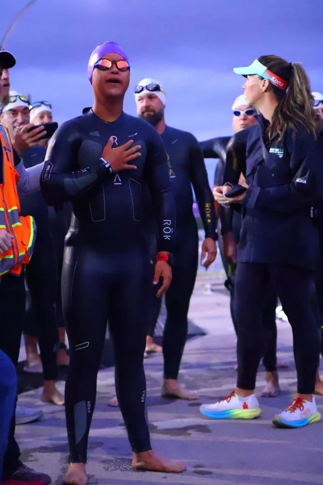 Group of athletes at a swimming event, with one athlete in the foreground holding her hand over her heart and wearing a black wetsuit and swimming cap.