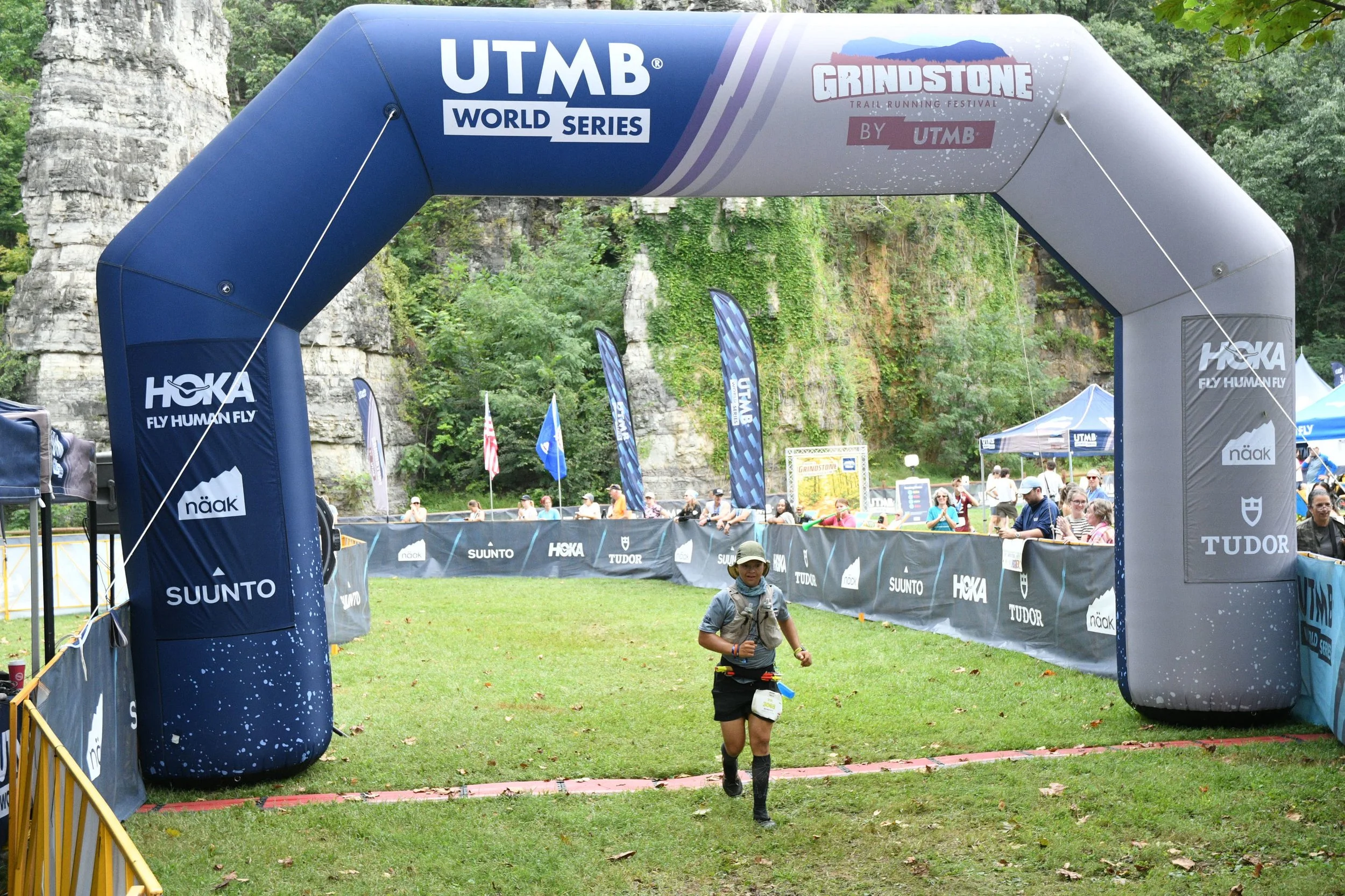 A person running at an outdoor trail running event, with a large blue balloon archway labeled 'UTMB World Series,' 'Grindstone,' and supported by banners and tents featuring sponsor logos. The scene is set in a natural area with tall cliffs and green trees in the background.