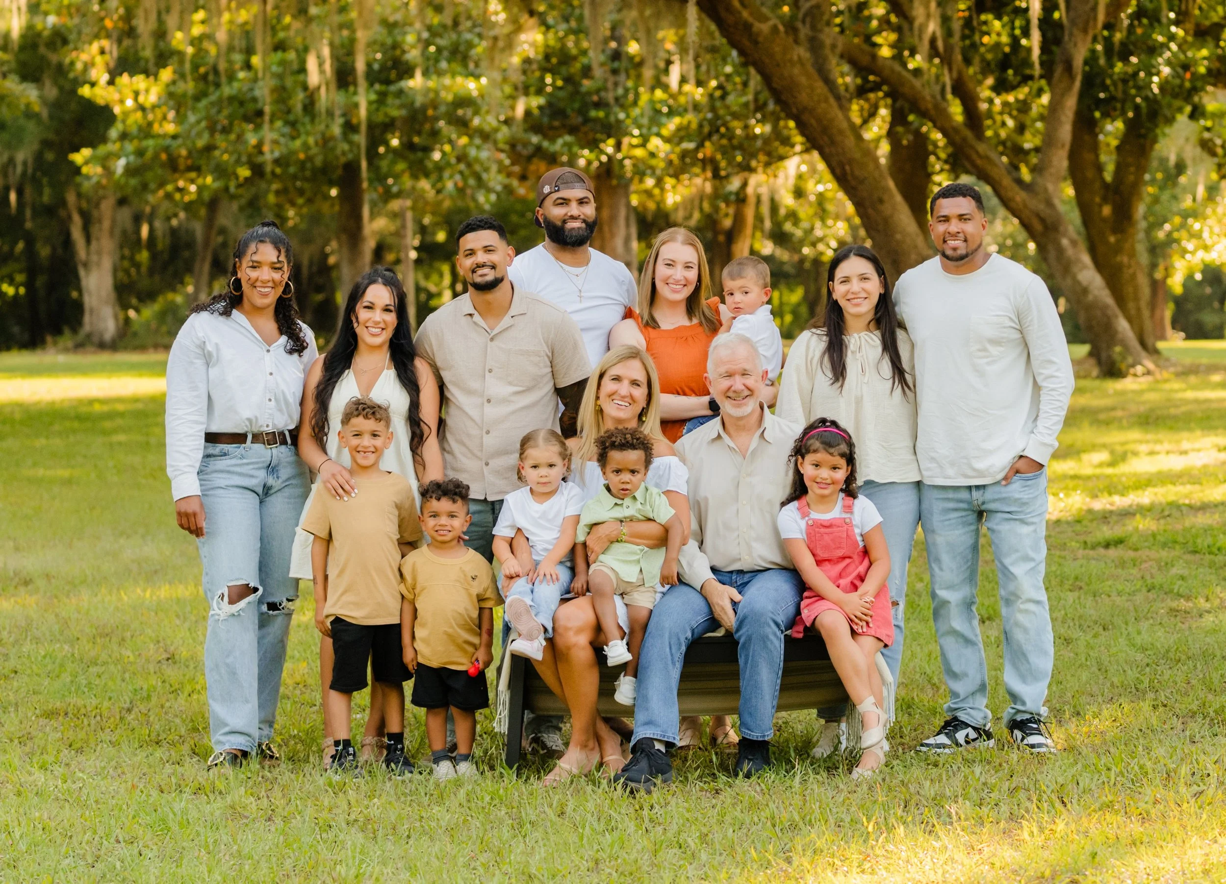 A large, multigenerational family of fifteen people posing outdoors in a park with trees and sunlight in the background. The family includes children, teenagers, and adults, all smiling and dressed casually.