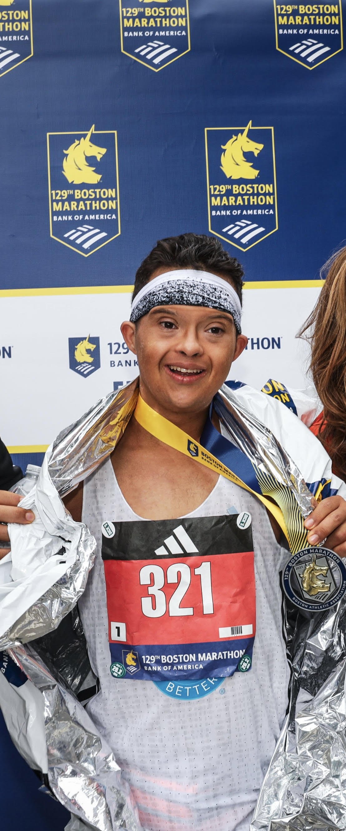A male marathon runner wearing a white tank top with bib number 321, smiling and holding a silver space blanket around his shoulders, with medals around his neck, celebrating his finish at the Boston Marathon. Behind him is a blue backdrop with the marathon's logo, a unicorn, and the text '129th Boston Marathon Bank of America'.