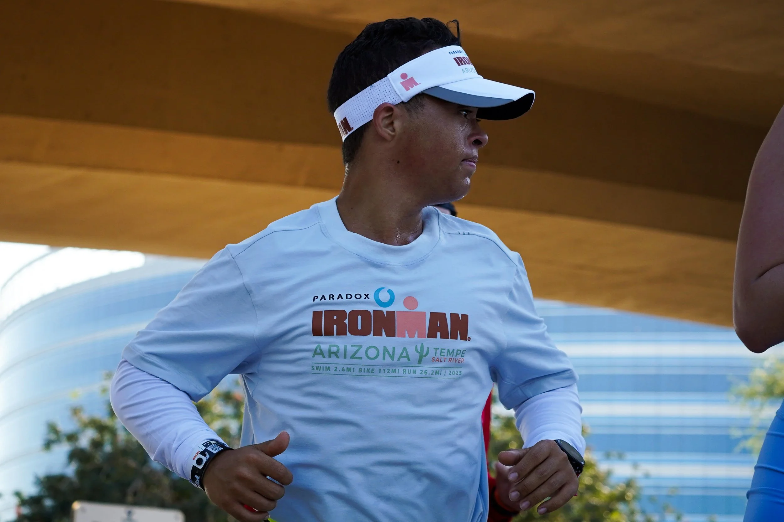 A man participating in the Ironman Arizona race, wearing a white long-sleeve shirt with race details and a white visor, standing outdoors with a modern building in the background.
