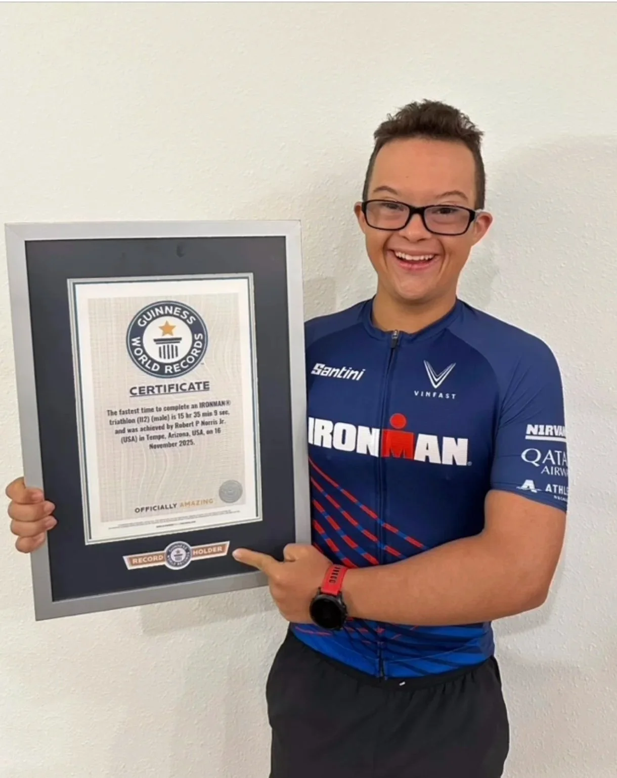 Young man smiling, wearing glasses and a blue Ironman triathlon shirt, holding a Guinness World Records certificate for the fastest time to complete an Ironman triathlon.
