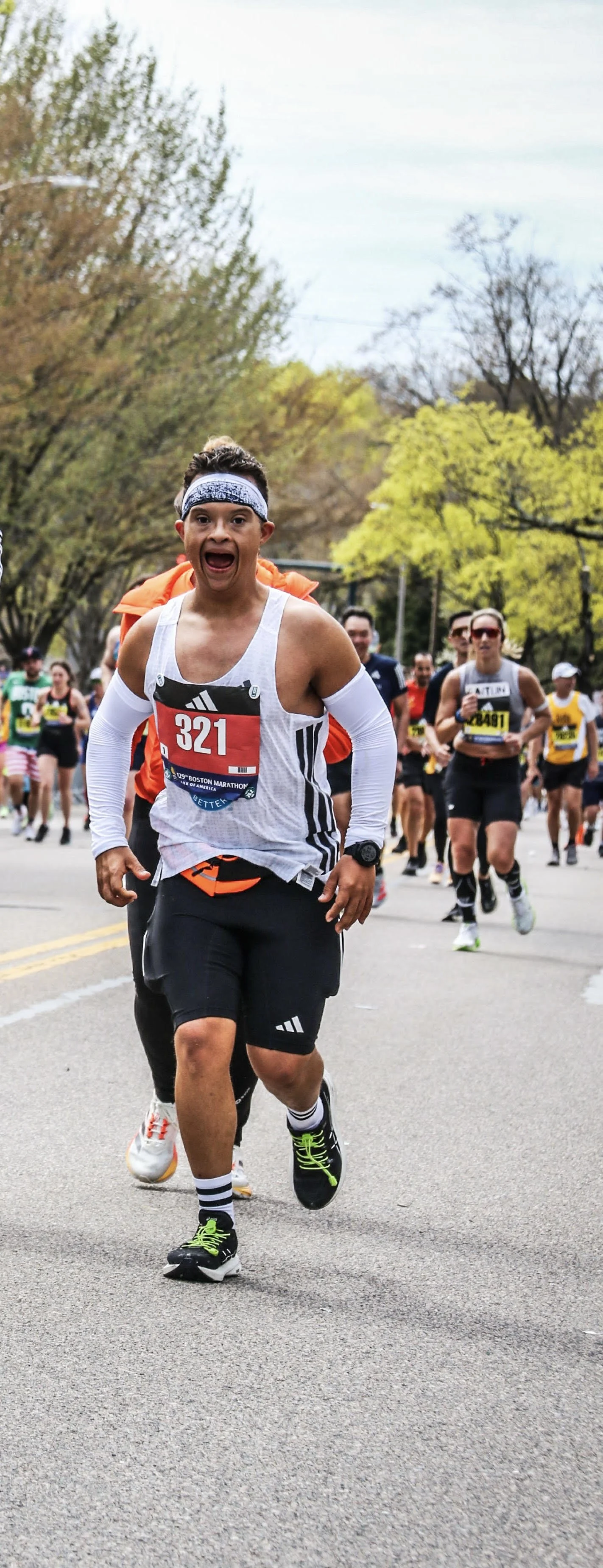 A group of runners participating in a marathon, with a young man in the foreground wearing a white tank top, black shorts, colorful shoes, and a headband, running on a street lined with trees.