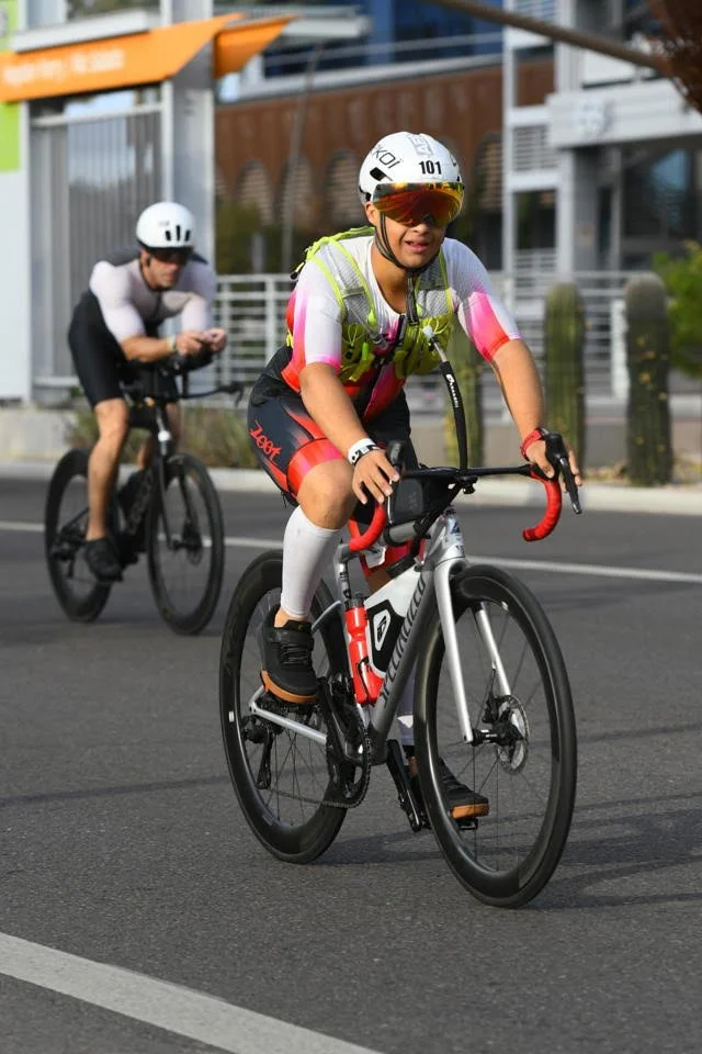 A young boy wearing a helmet, sunglasses, and cycling gear is riding a road bike on the street. In the background, another person on a bike is visible, along with city buildings and a fenced area.