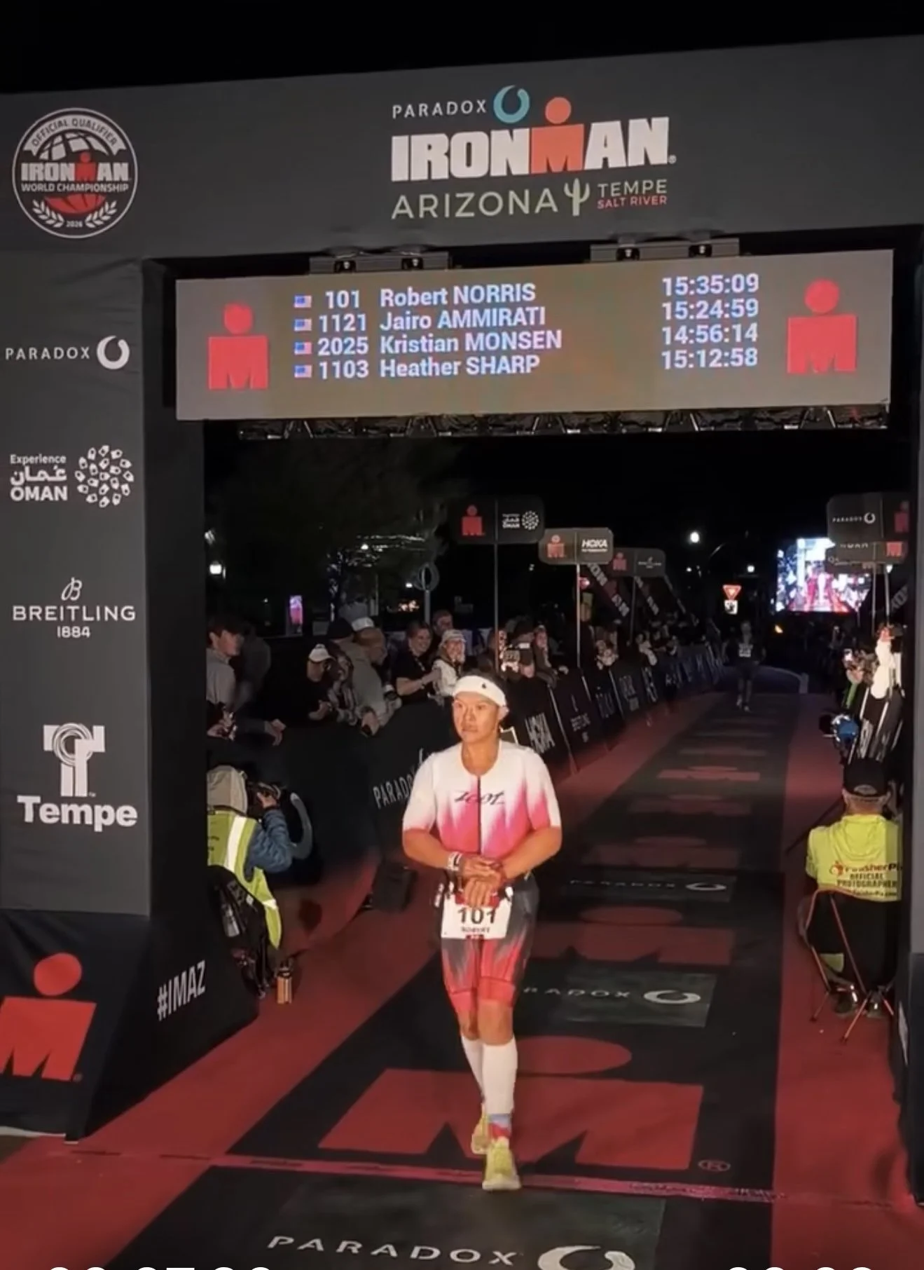 Ironman triathlon finish line in Tempe, Arizona with athlete crossing, race information board displaying names and times, and spectators in the background.