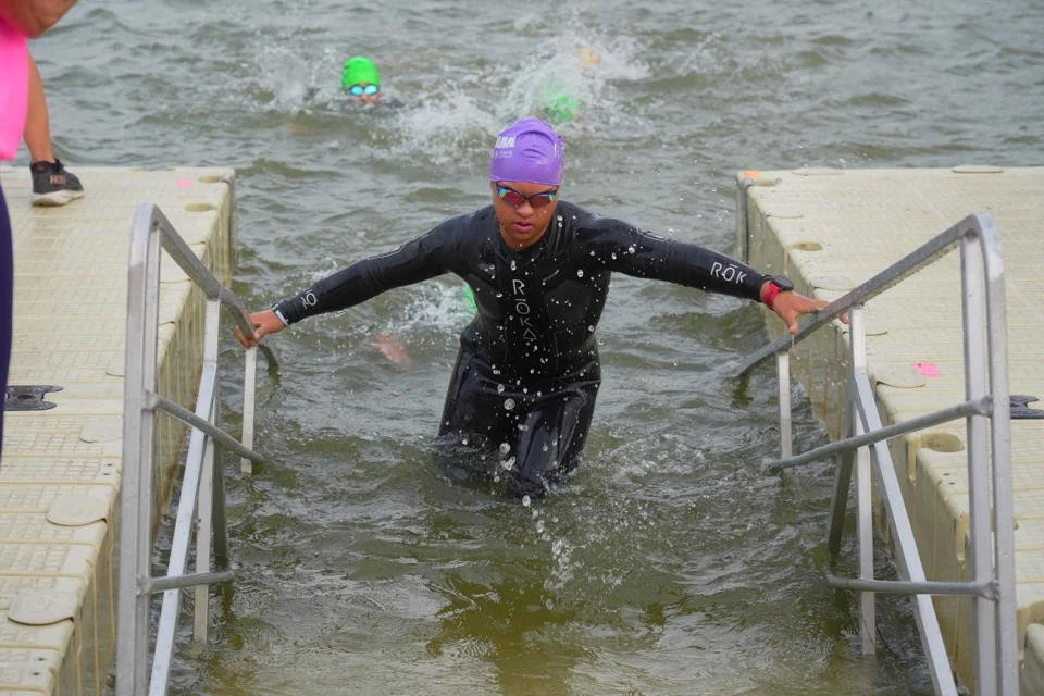 A swimmer wearing a purple swim cap, goggles, and a black wetsuit is emerging from the water onto a floating dock, using metal handrails for support.