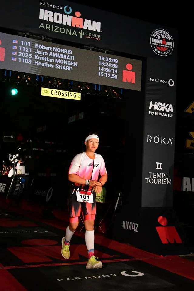 Female athlete crossing the finish line at the Ironman Arizona race in Tempe, salt River, with a digital scoreboard above displaying her and other participants' names and finishing times.
