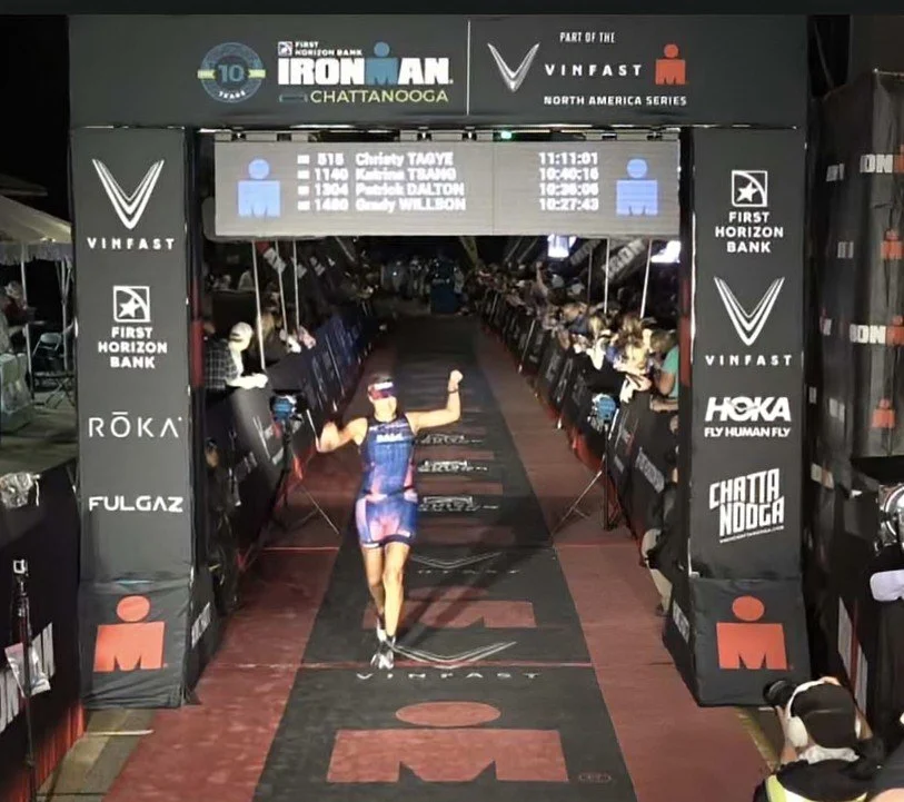 A female athlete crosses the finish line of an Ironman race in Chattanooga, running on a designated path with sponsors' logos. She celebrates with raised arms, and spectators are on both sides of the course. An electronic timer displays race times and the athlete's name, Charity TAYE.