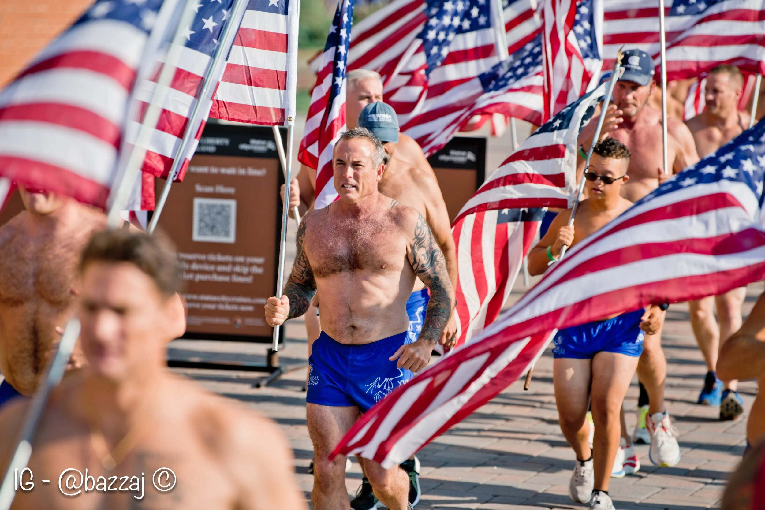 Group of diverse shirtless men and a woman running outdoors with American flags, participating in a patriotic event or race.