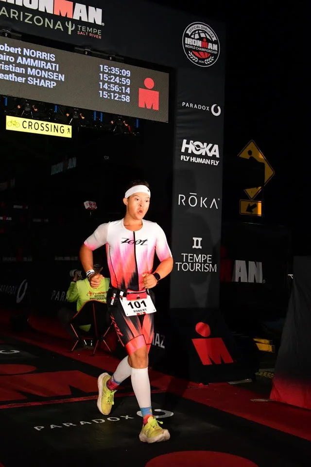 A male athlete wearing a white and pink running outfit with a race number 101 is running in an indoor race event. The background has digital timers displaying times, various sponsor logos, and signs including 'Crossing' and directional arrows.
