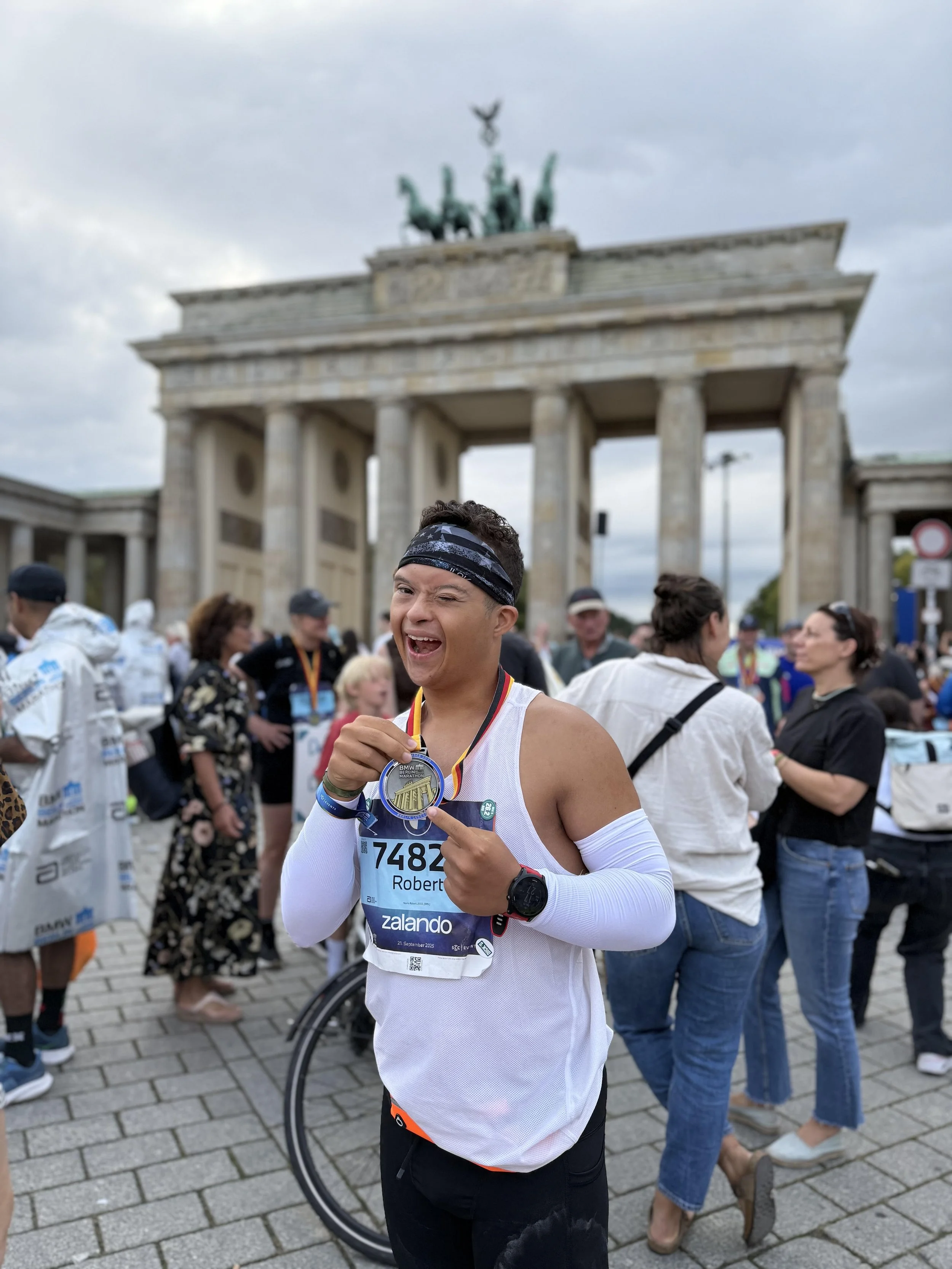 A young man smiling and holding a medal in front of the Brandenburg Gate in Berlin, Germany during a marathon event, with other runners and spectators around him.