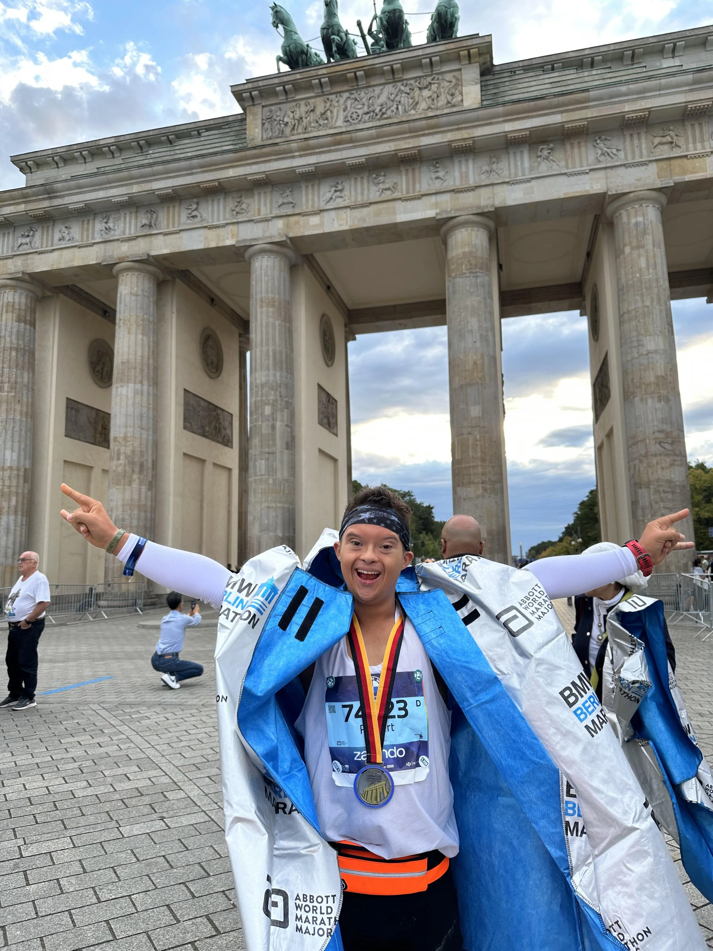 A young marathon finisher wearing a medal, holding his arms up in celebration, standing in front of the Brandenburg Gate in Berlin. He is smiling widely, dressed in running gear and a race bib.