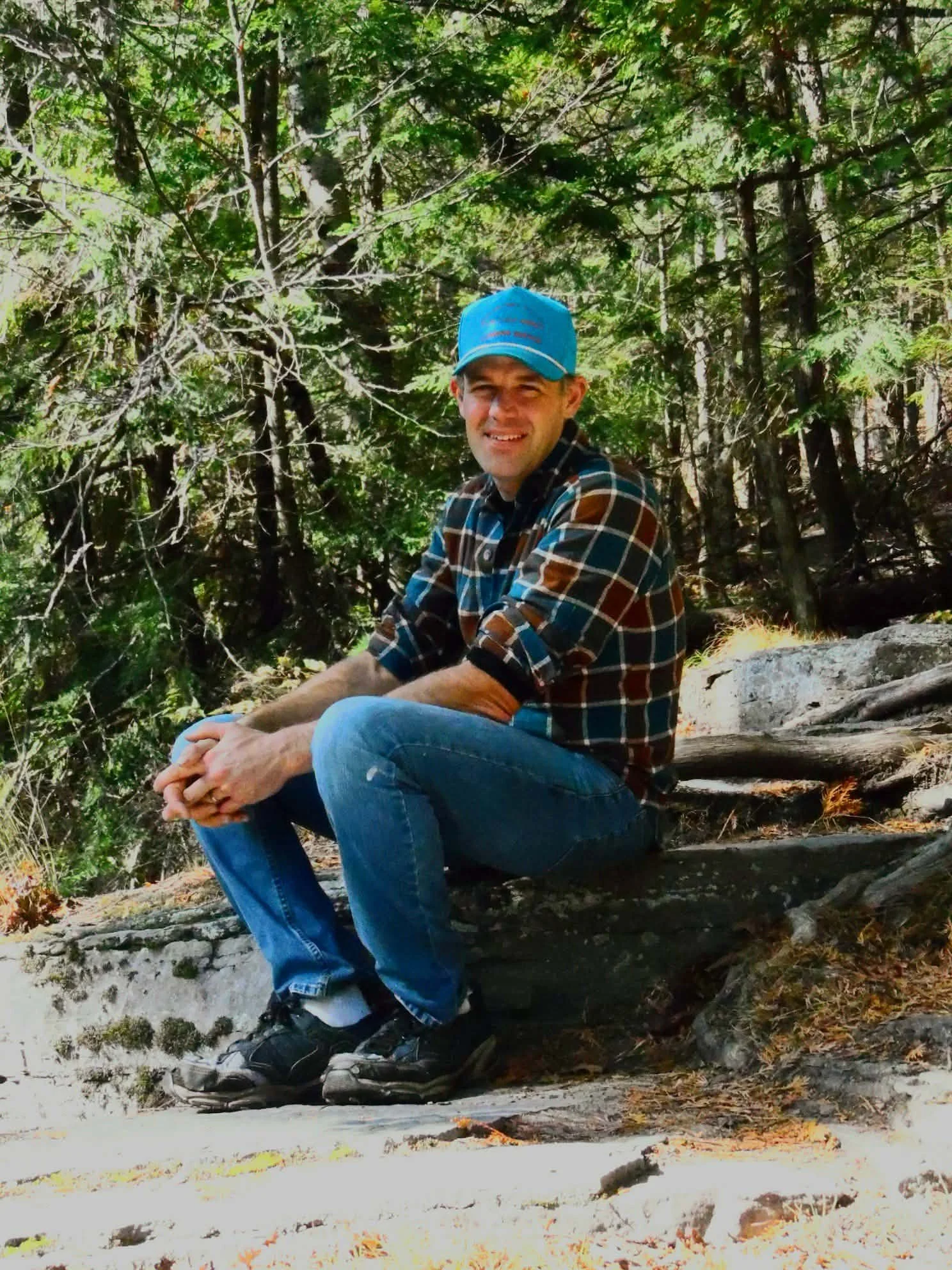 A man sitting on a flat rock in a forest, wearing a blue baseball cap, a plaid shirt, jeans, and hiking shoes, surrounded by trees and greenery.