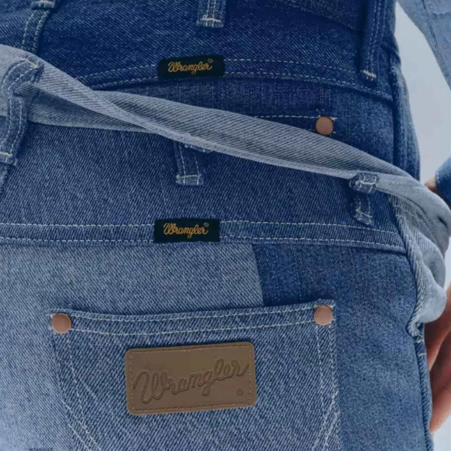 Close-up of denim jeans with a Wrangler logo patch and buttons, worn by a person, with the waistband folded over.