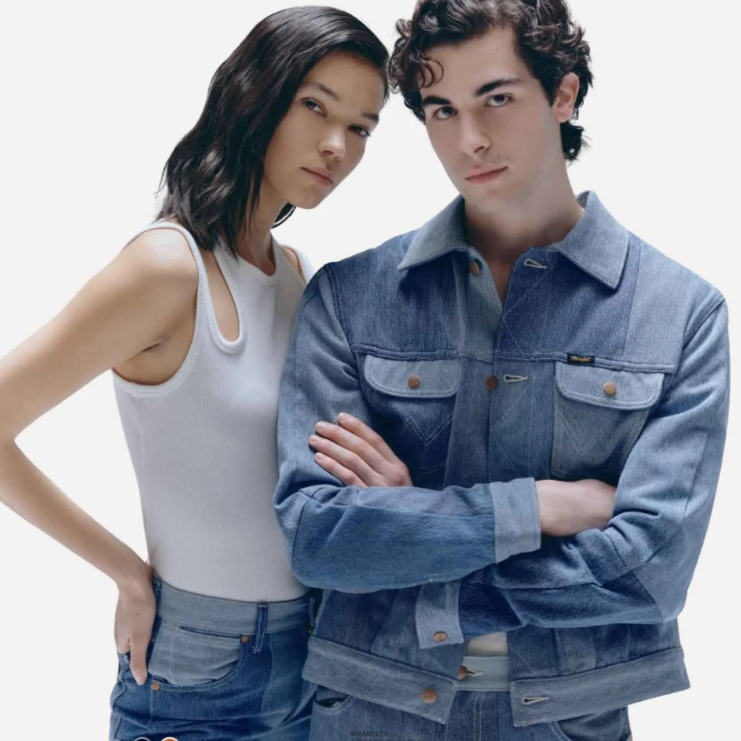 A young man and woman standing close together with serious expressions, the woman has dark hair and wears a white tank top, the man has curly hair and wears a denim jacket, both with arms crossed.