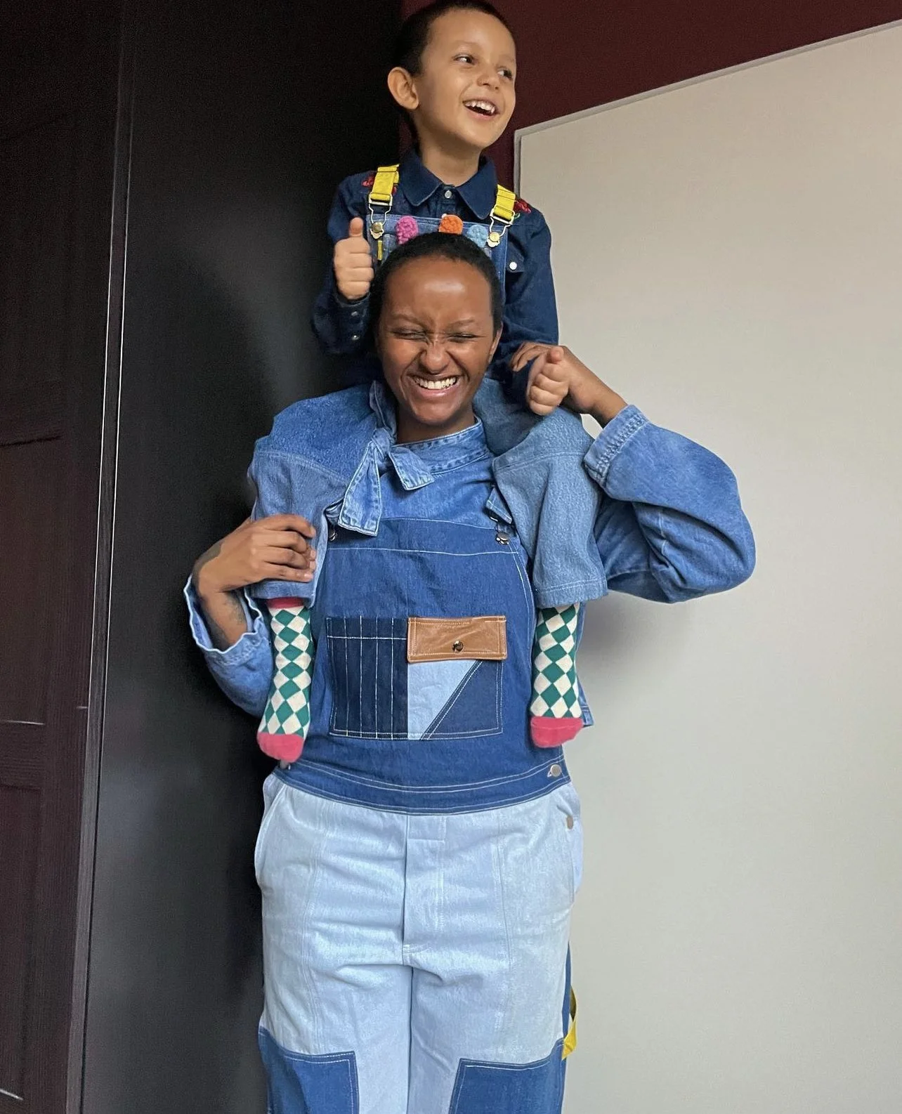 A woman carrying a young girl on her shoulders, both smiling happily indoors and both wearing upcycled fashion designs from Beyond Remade.