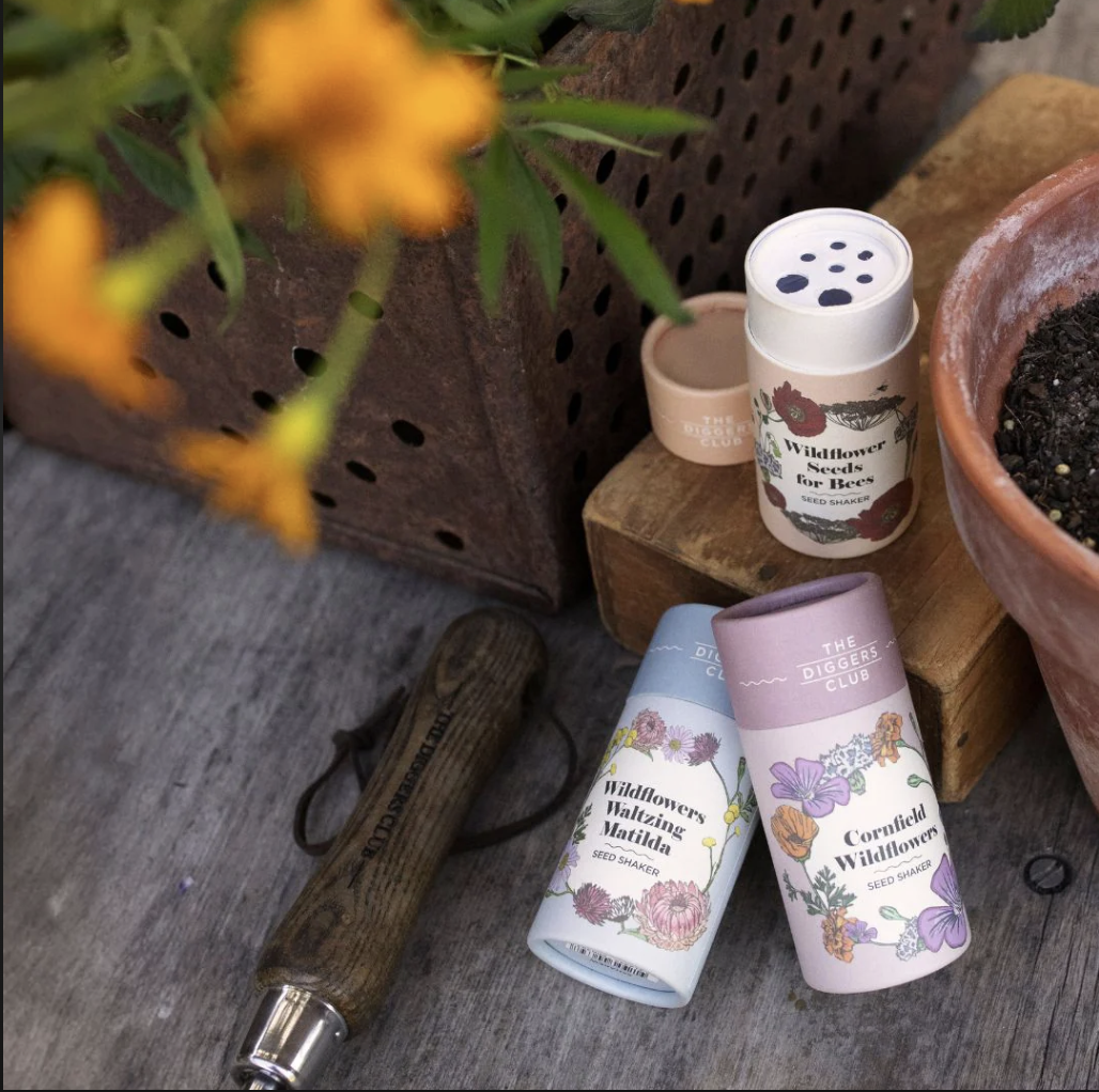Seed shakers labeled 'Wildflower Seeds for Bees,' 'Wildflowers Wandering Matilda,' and 'Cornfield Wildflowers,' next to a wooden-handled gardening tool and a clay pot with soil, on a wooden surface.