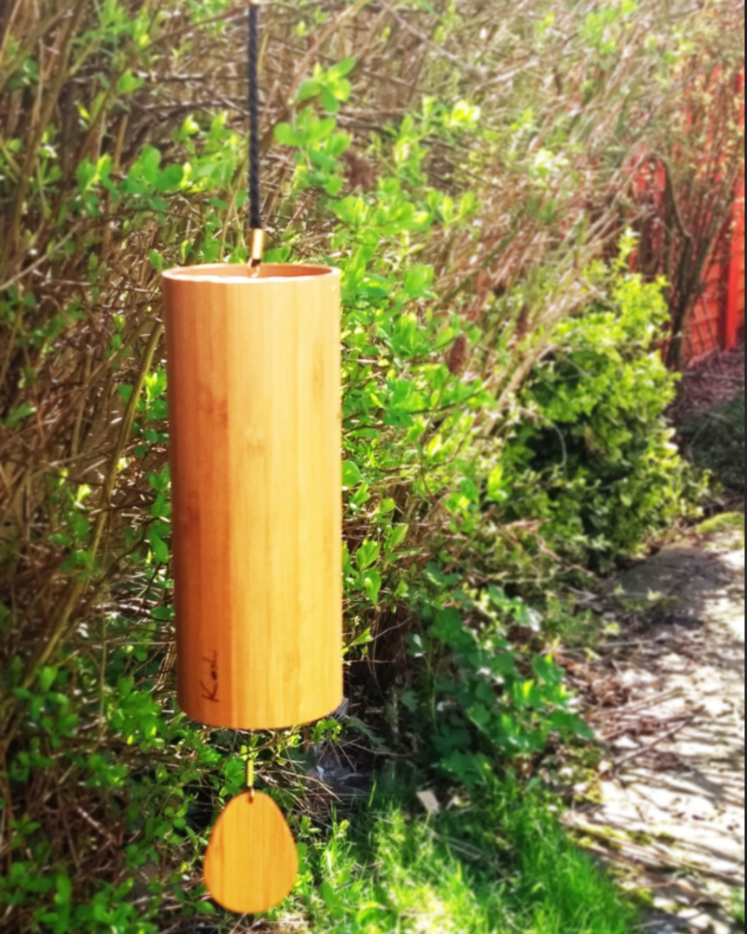 Wooden wind chime hanging from a branch in a garden with green bushes and a red fence in the background.