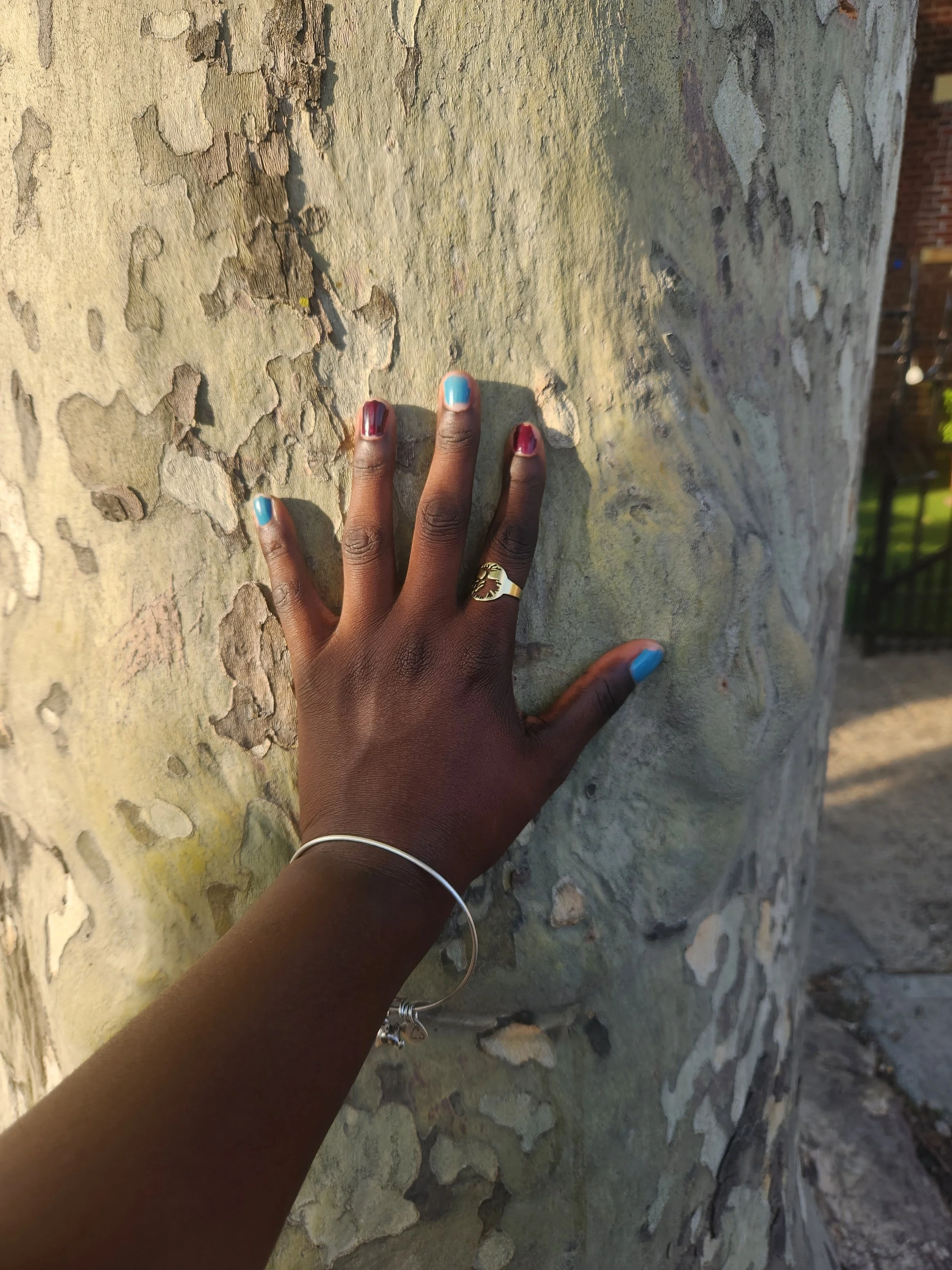 A person's hand with polished nails in blue and pink resting on a textured, light-colored stone wall, wearing a silver bracelet, a silver ring with a gold design, and a silver ring with a star charm, with a background of trees and a brick building.