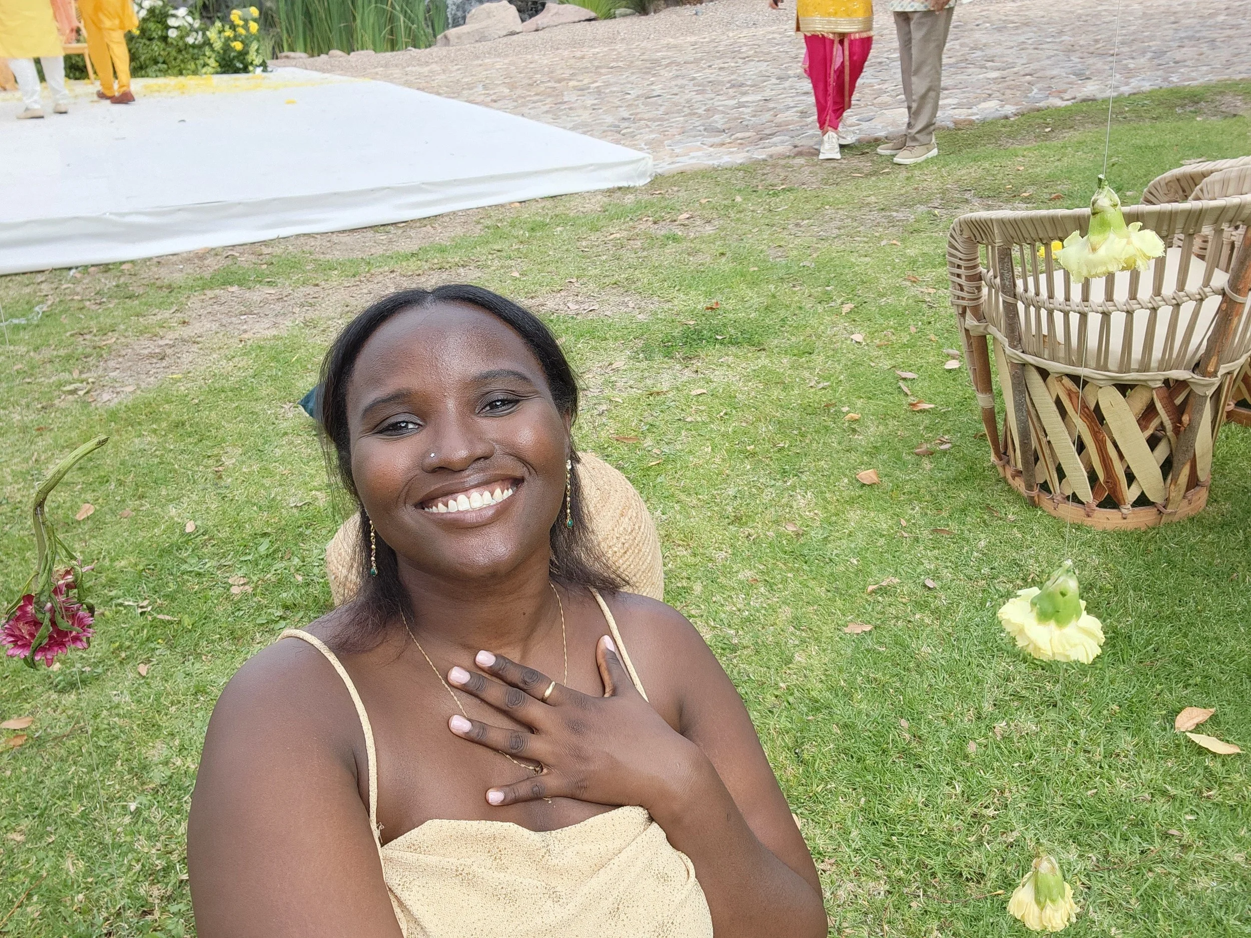 Smiling woman with hand on chest sitting on grass at outdoor event, with hanging yellow flowers and a wicker chair in the background.